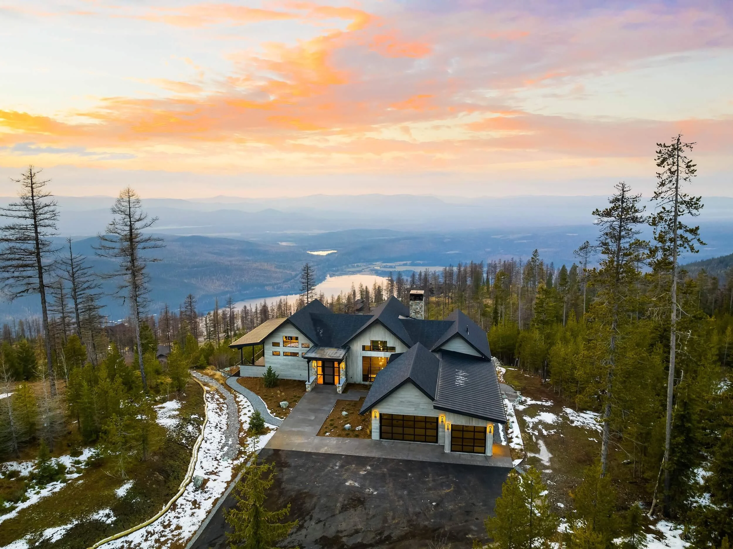 A modern house with a gray roof and large windows is situated in a wooded area with tall trees, some of which are bare. The house features a driveway and garage, and a stone pathway leads through the backyard. The landscape includes patches of snow, indicating a late winter or early spring setting. The background showcases distant mountains and a colorful sky with pastel pink and orange clouds during sunset.