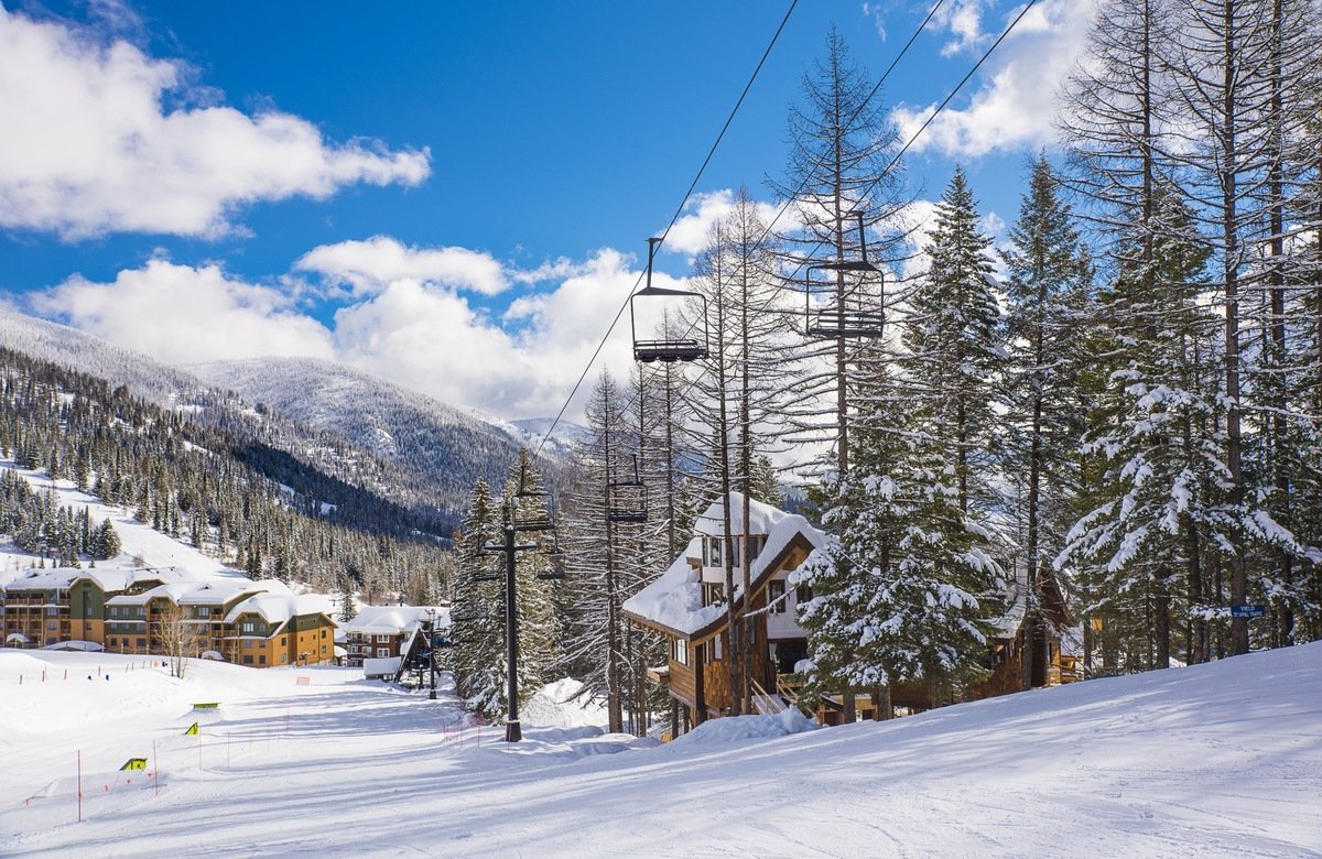 Snow-covered ski resort with a ski lift, pine trees, and buildings in the background under a partly cloudy sky.