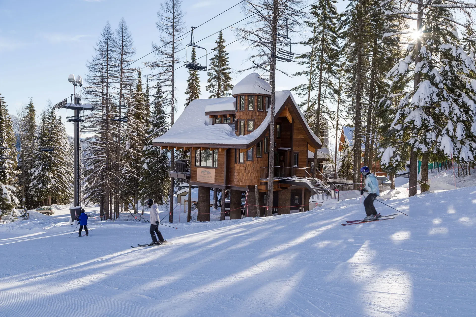 A snowy ski resort with a ski lift, wooden building, and skiers on the slopes surrounded by tall snow-covered trees, with the sun shining through the branches.