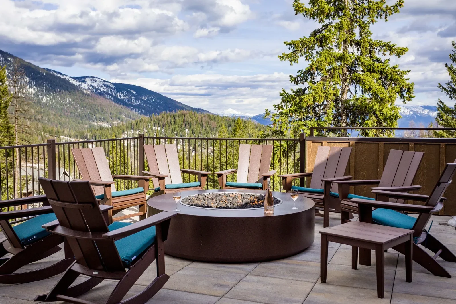 Outdoor patio with wooden chairs around a fire pit overlooking a mountainous forested landscape.