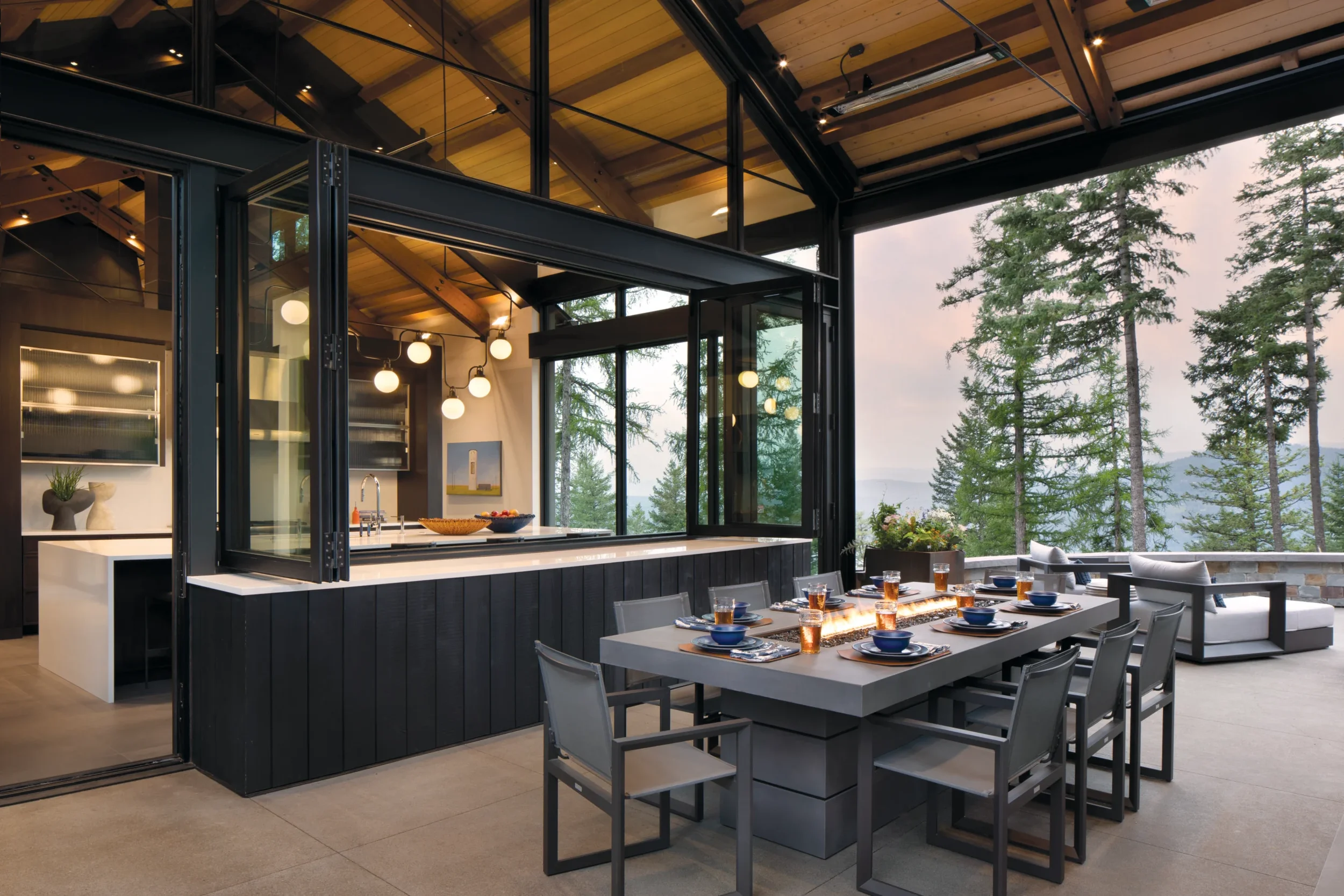 Modern outdoor dining area with a table set with plates and glasses in front of a glass-enclosed kitchen, surrounded by outdoor trees and a mountainous landscape in the background.