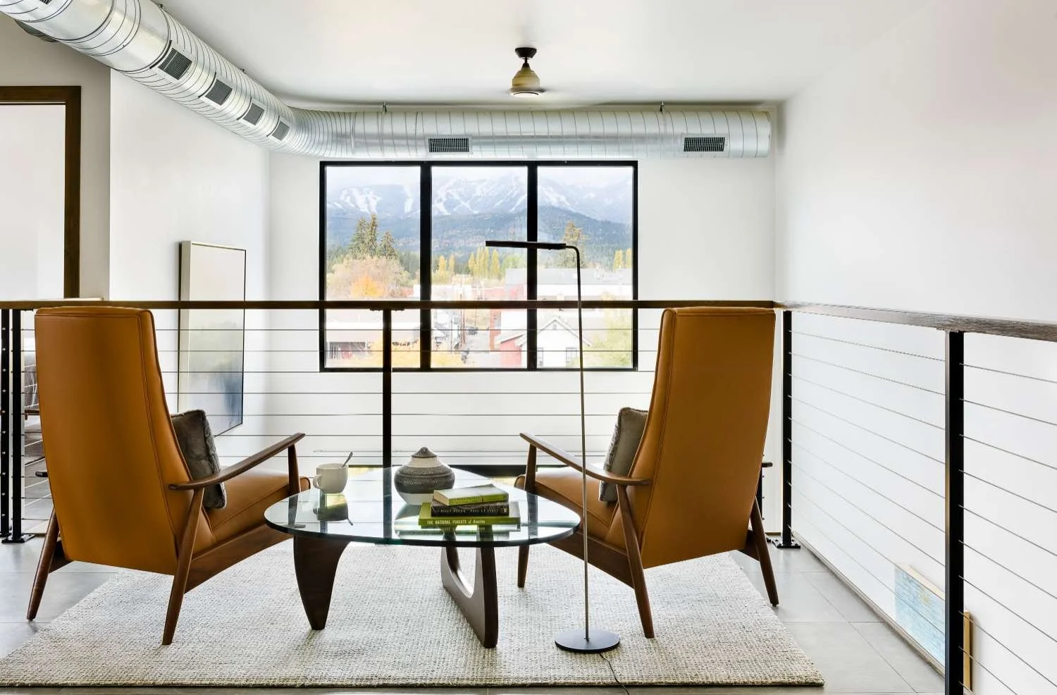 A modern sitting area with two yellow leather armchairs, a glass coffee table with books and a teacup, a tall floor lamp, and a large window with scenic mountain view. Industrial ductwork runs across the ceiling.