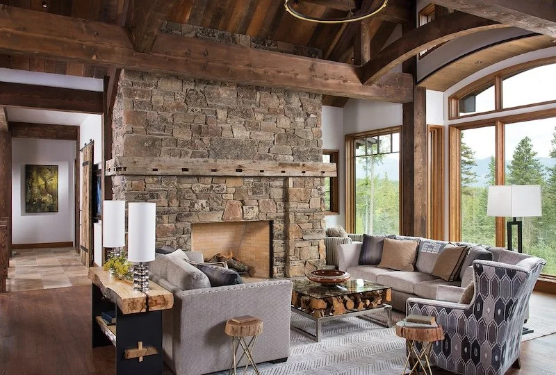 Living room with large stone fireplace, wood ceiling beams, and big windows showing trees outside, furnished with sofas, armchair, coffee table, and lamps.