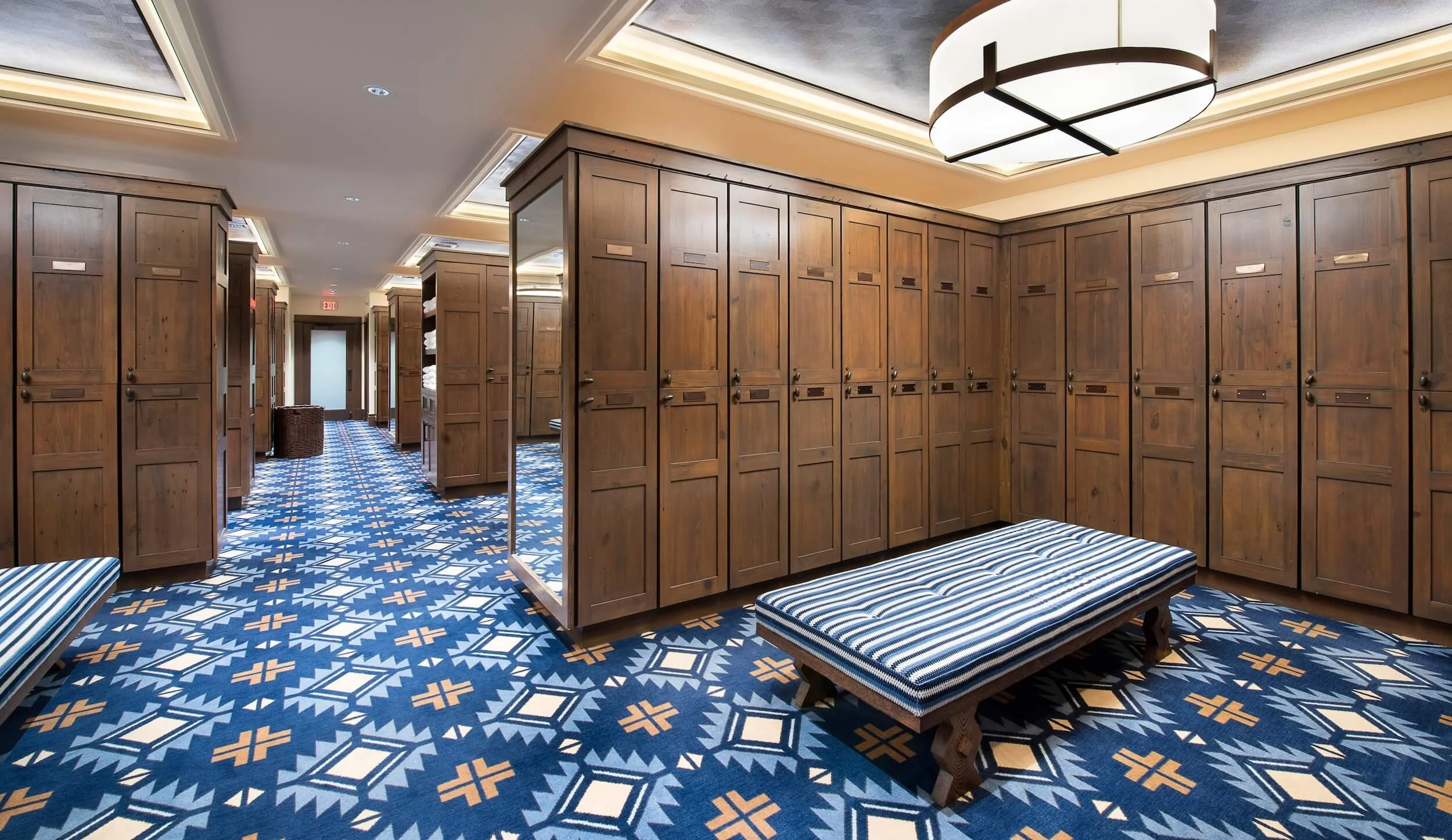 A locker room with wooden lockers lining the walls, striped benches, and a patterned blue and yellow carpet. The ceiling has recessed lighting and a large modern ceiling light fixture.