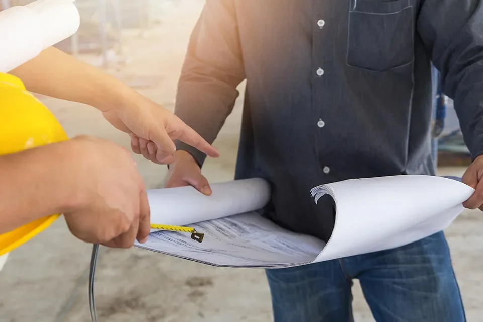 Two individuals examining a large rolled-up set of architectural blueprints or plans at a construction site.