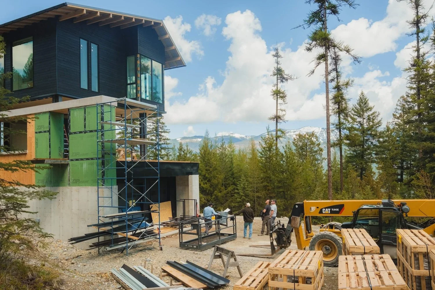 Construction site of a modern multi-story house in a forested area with mountains in the background. Workers are present, and building materials, scaffolding, and construction equipment are visible.