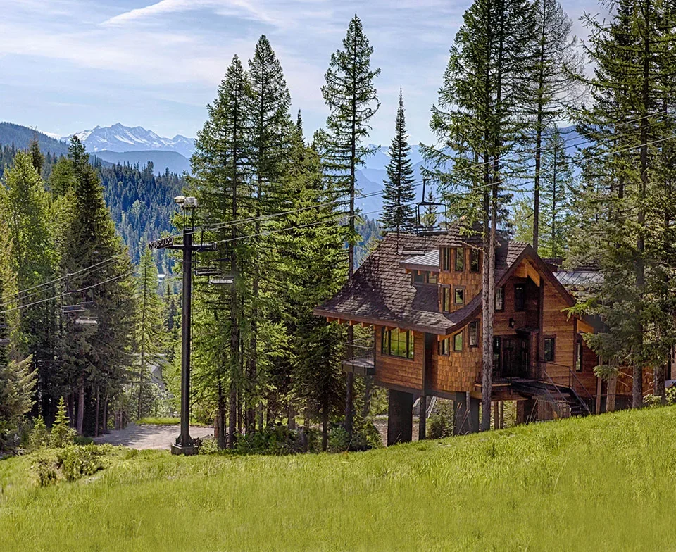 A wooden house surrounded by tall pine trees with a mountain range in the background.