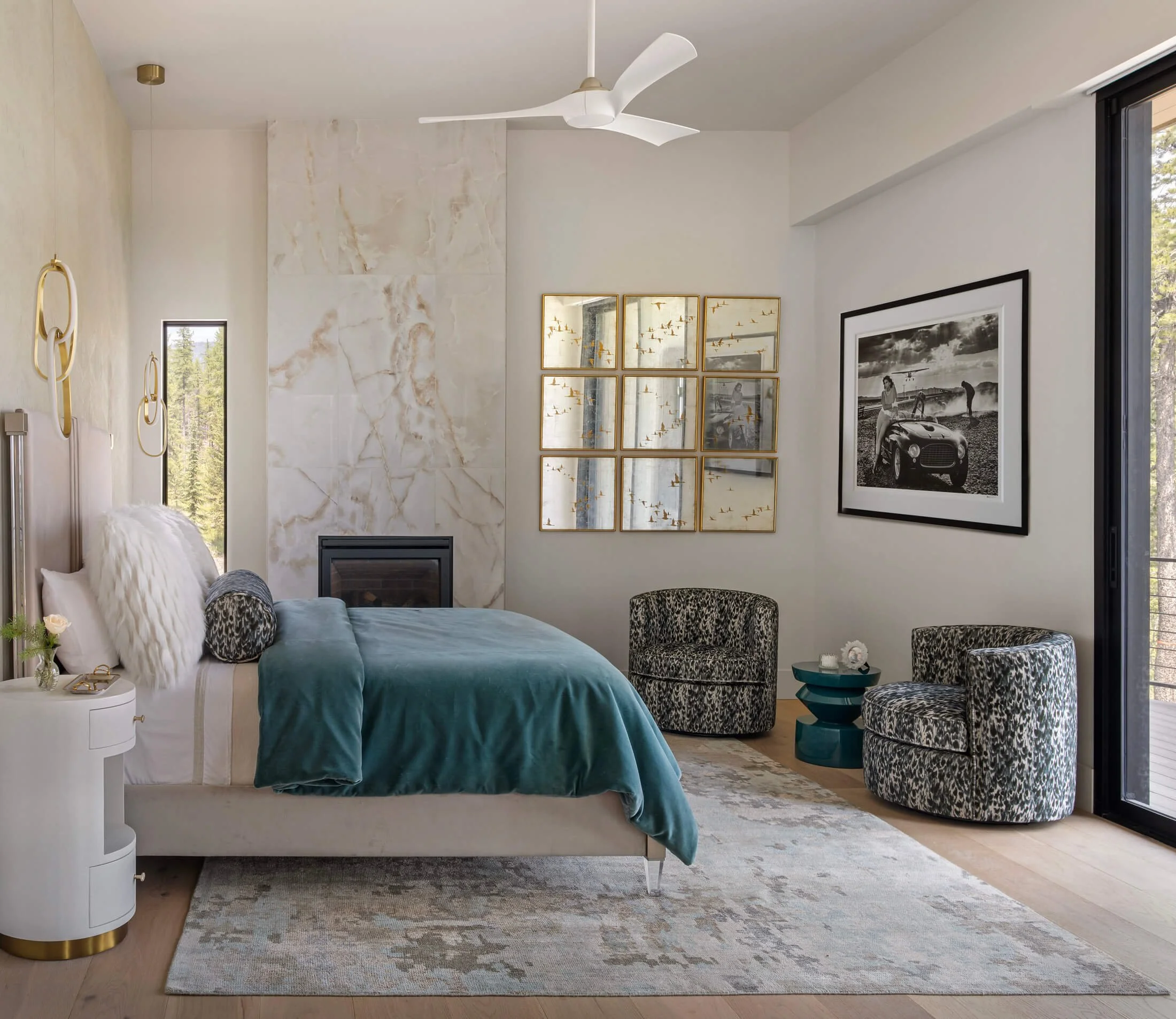 A modern bedroom with a white ceiling fan, framed photographs on the wall, a large window, a teal bed with fluffy pillows, and patterned chairs next to a small teal side table.