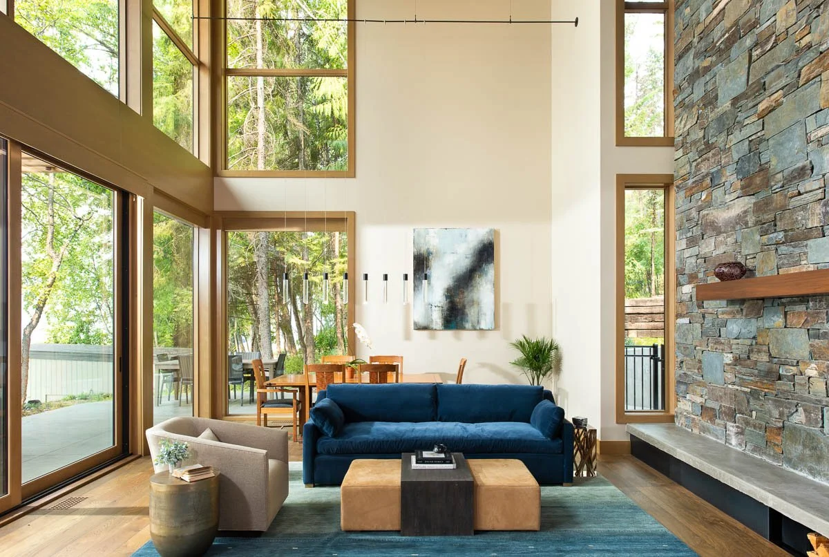 Modern living room with large windows, a blue velvet sofa, beige armchair, wooden coffee table, stone fireplace, and abstract art on the wall.