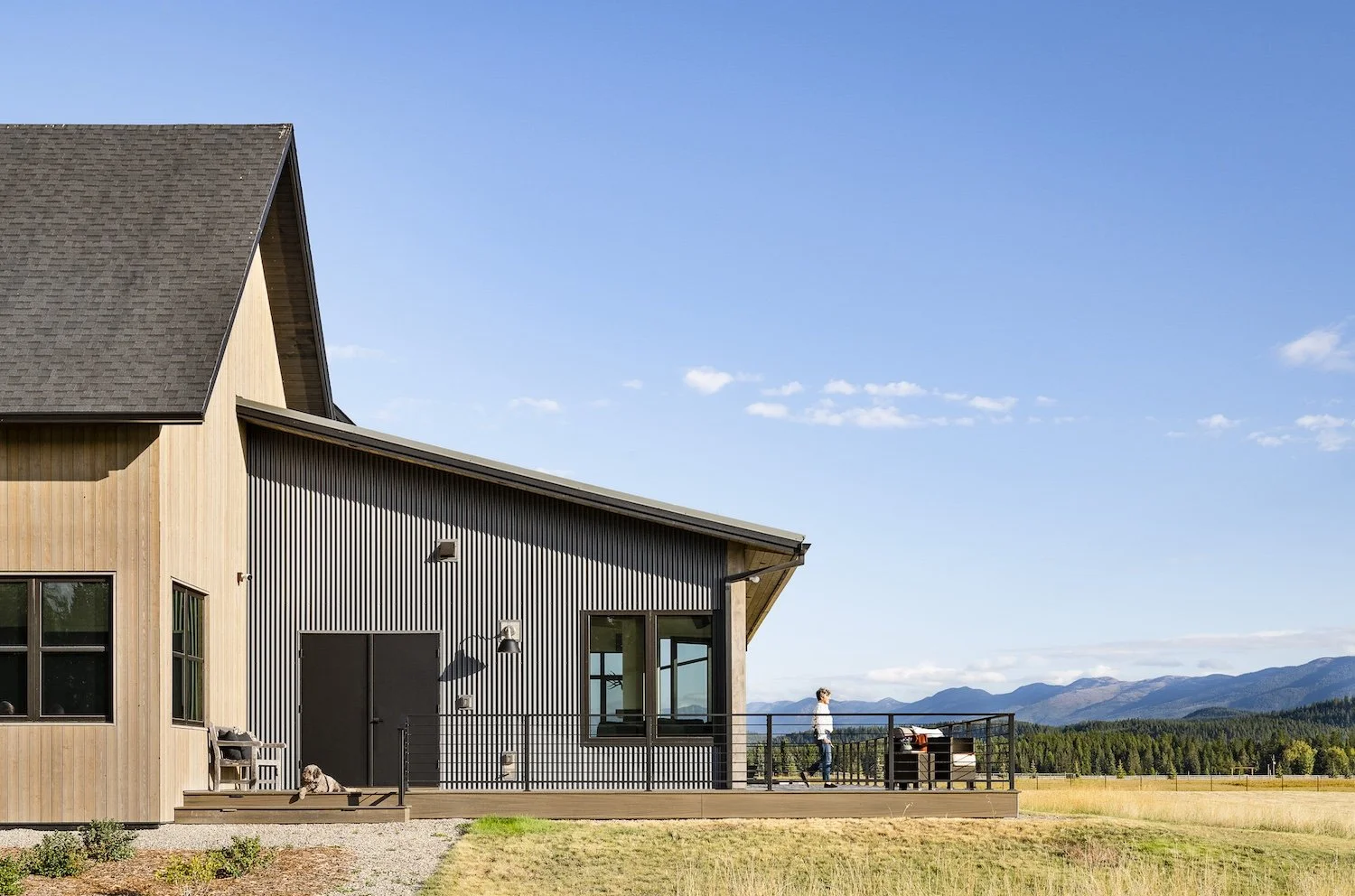 A modern house with a combination of wood and metal exterior, a deck with a person standing on it, and distant mountains under a blue sky.