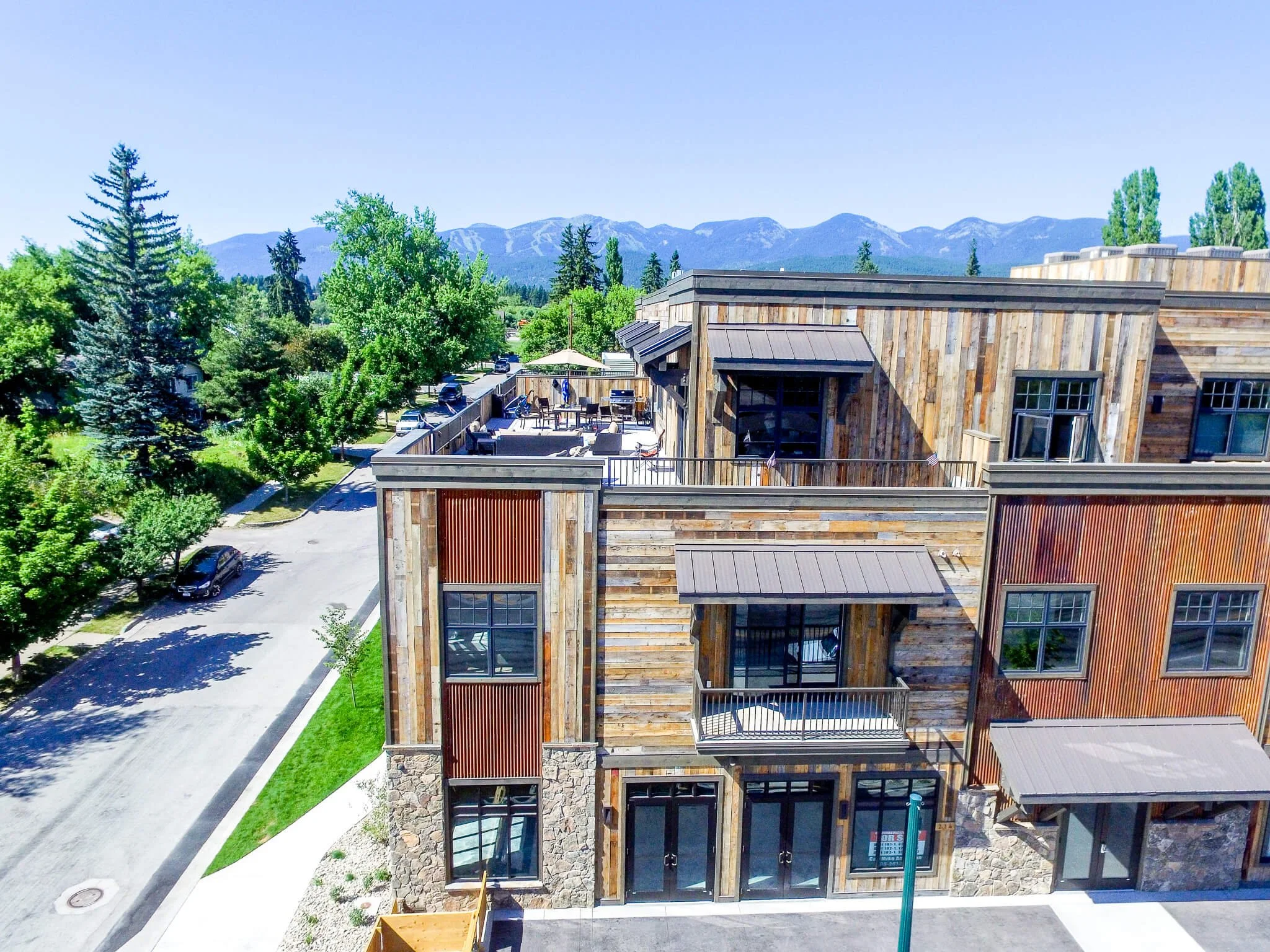 A multi-story building with a rustic wooden exterior, large windows, and a balcony. The building is in a green area with trees, a parking lot, and mountain ranges in the background under a blue sky.