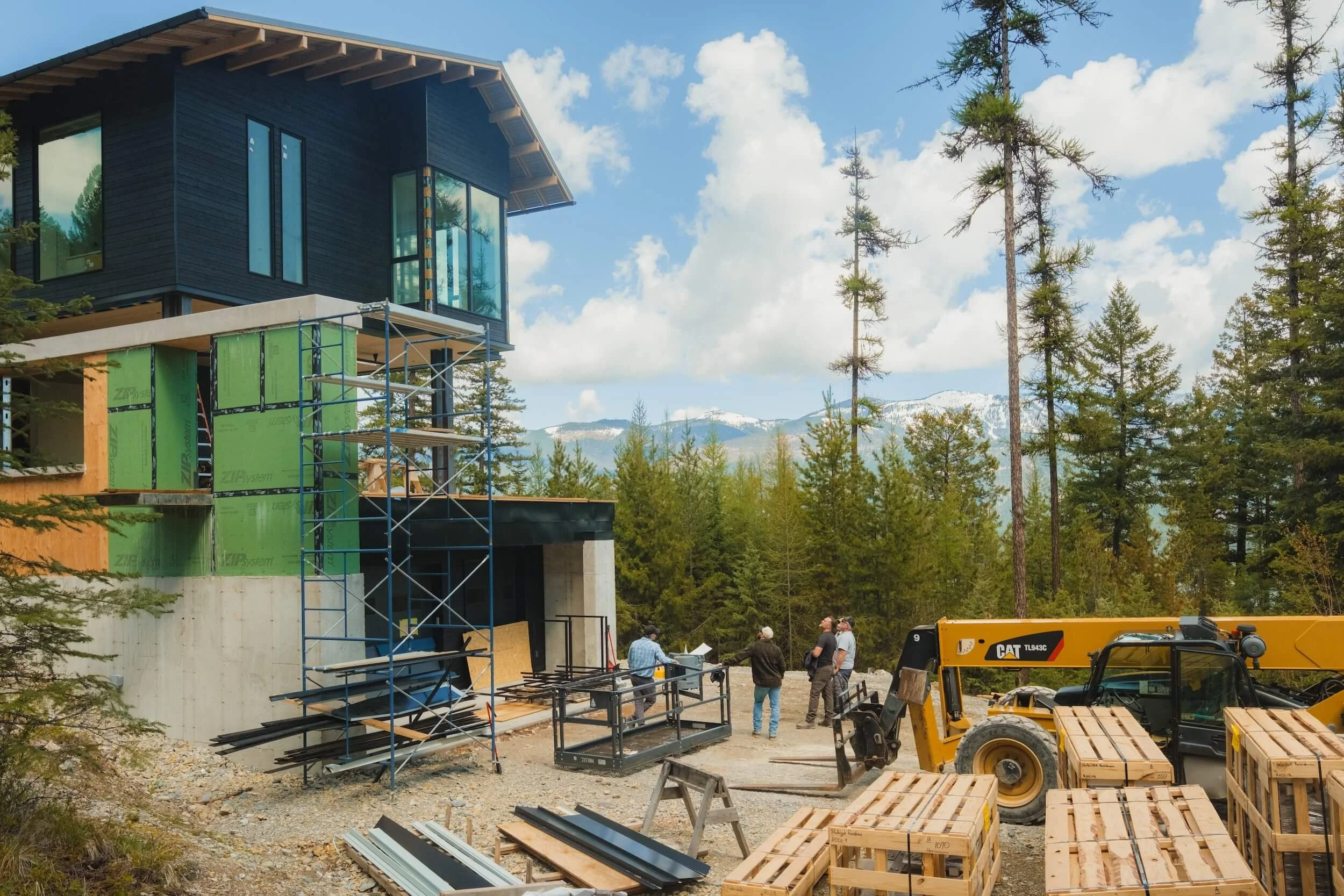 Construction site of a modern black house with large windows, surrounded by trees and mountains in the background, with workers and construction equipment present.