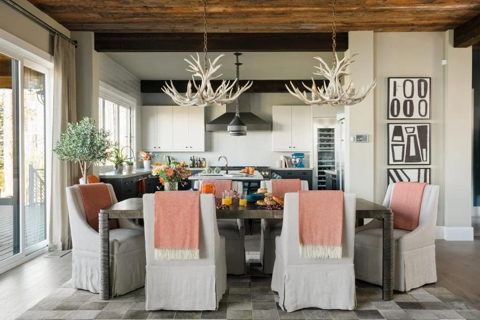 Dining area with a dark wood table, eight chairs with beige slipcovers and pink throws, and antler chandelier, open kitchen with white cabinets, black accents, and a sliding glass door leading outside.