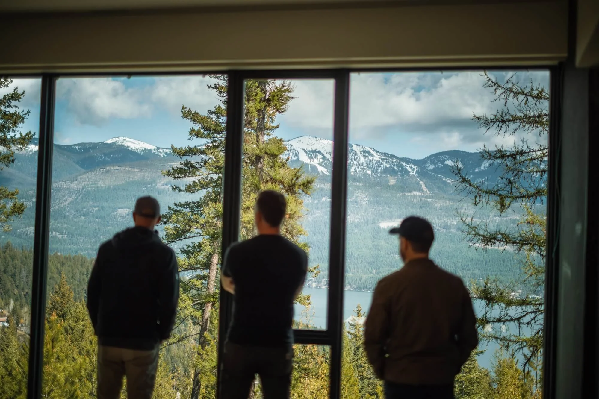 Four people standing inside a building looking out a large window at a mountainous landscape with snow-capped peaks, trees, and a lake.