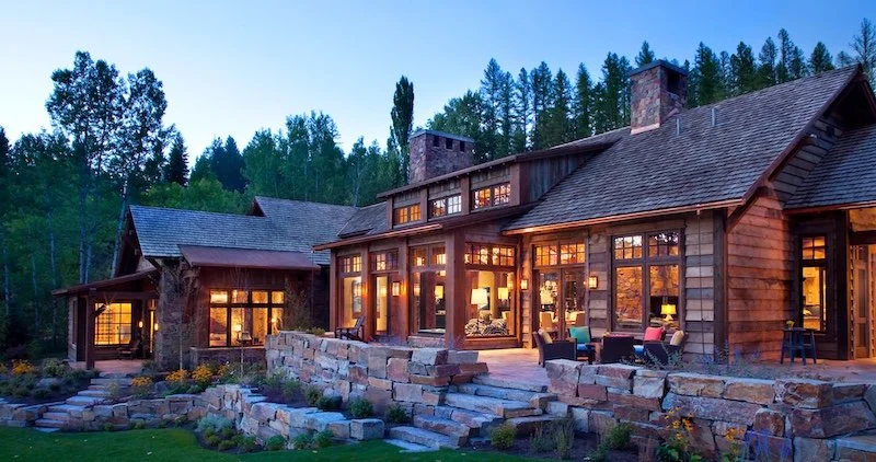 Large rustic wooden house with large windows and a stone patio, surrounded by trees, during evening with interior lights on.