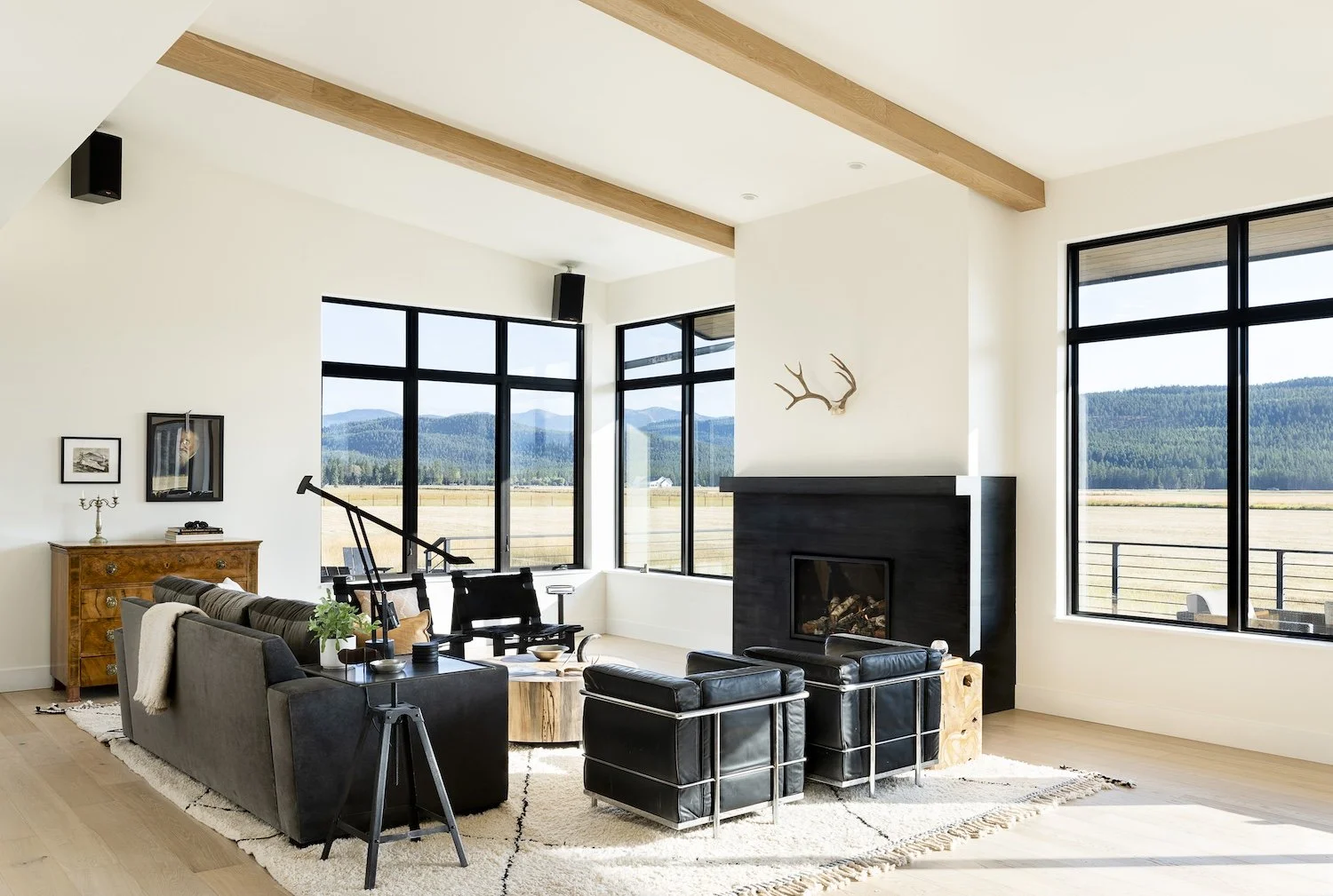Modern living room with large windows showing a mountain view, black fireplace, black leather and fabric furniture, wooden accents, and decorative items.