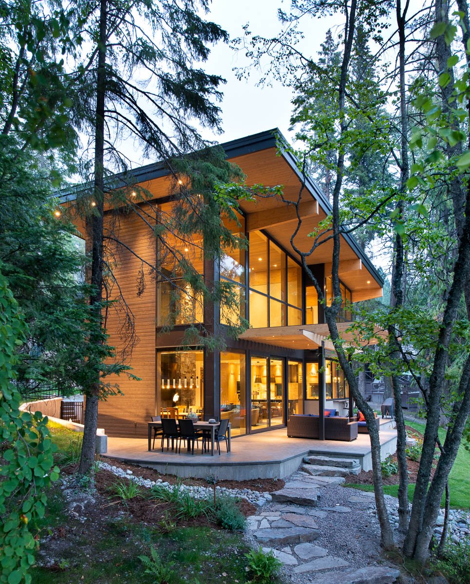 A modern two-story house with large glass windows, situated among trees in a wooded area. The house is illuminated from inside, and a patio with outdoor furniture is visible in front of the house.