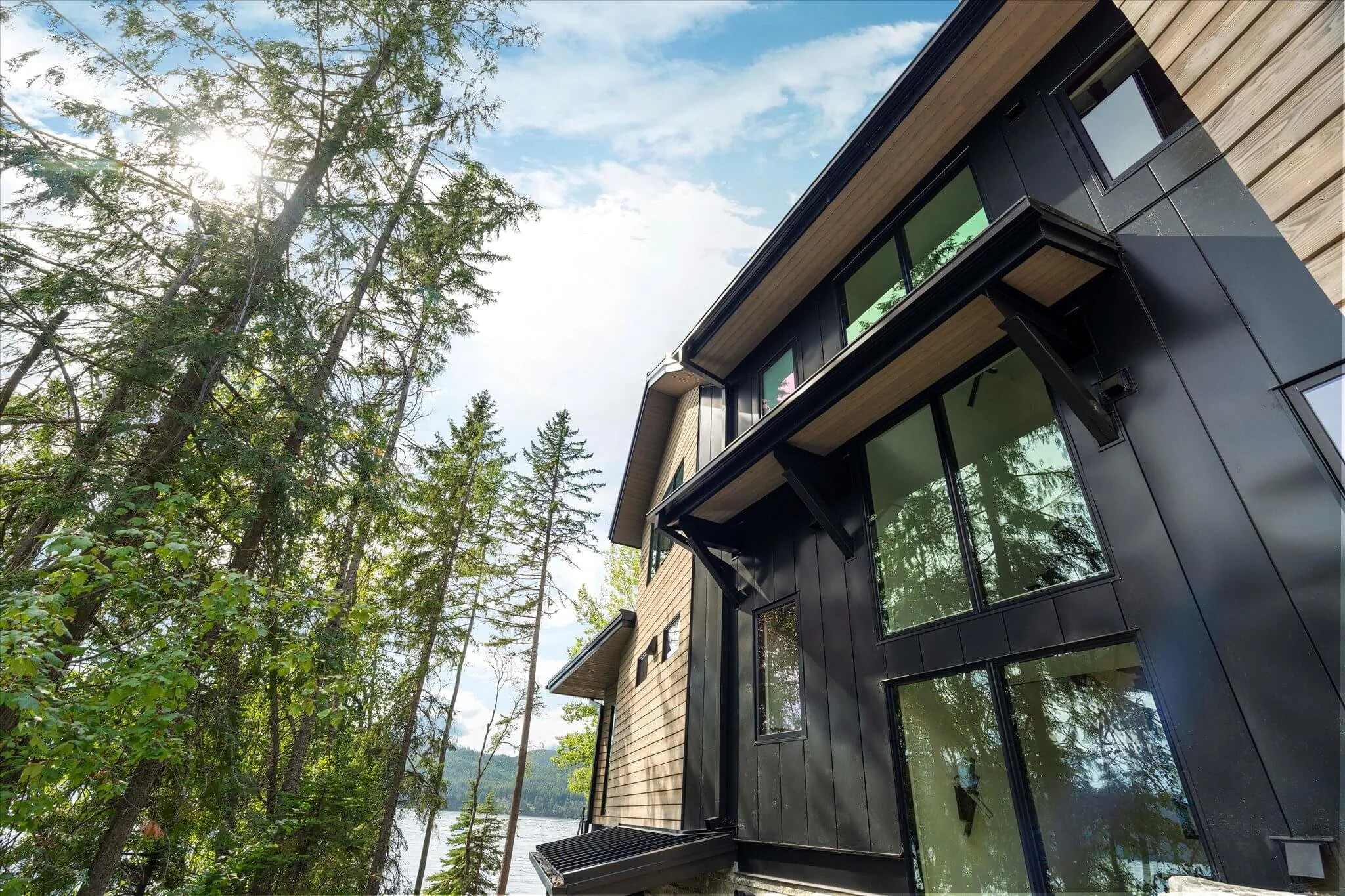 Modern multi-story house with large windows and black exterior, surrounded by tall green trees and overlooking a body of water under a partly cloudy sky.