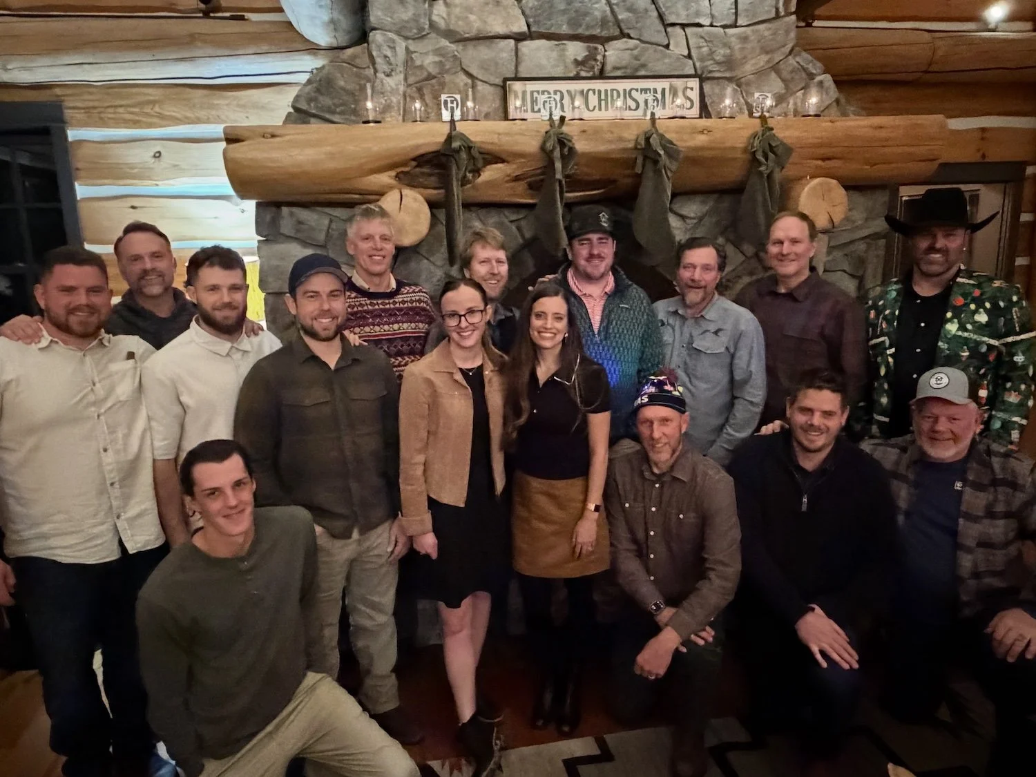 Group of people gathered in a cozy log cabin, standing in front of a stone fireplace with a wooden log mantel decorated with a sign that says 'Merry Christmas' and hanging stockings, celebrating the holiday season.