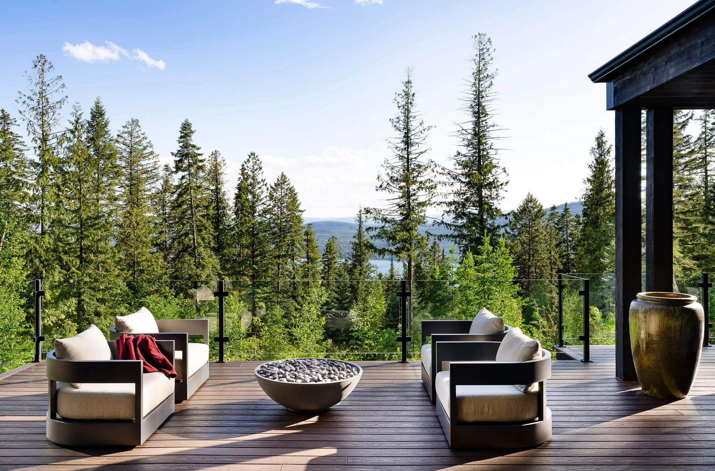 A modern outdoor patio with white cushioned chairs, a round fire pit filled with stones, and a scenic view of a forest and mountains in the background.