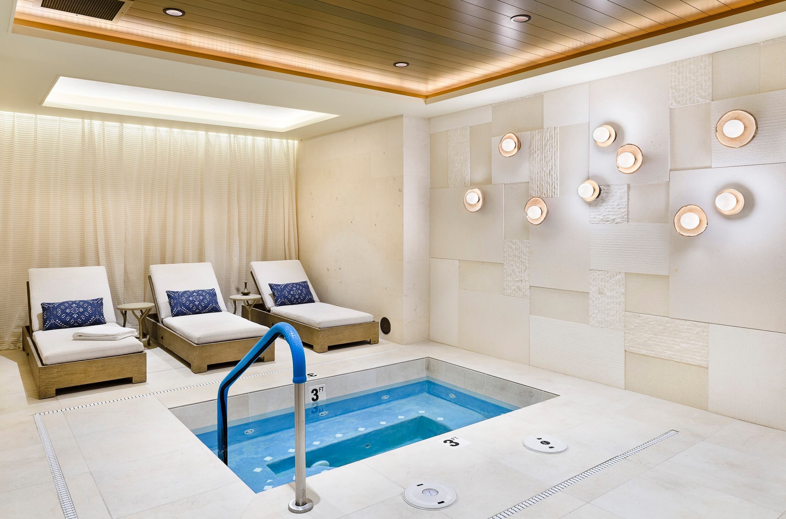 Indoor pool area with two lounge chairs with blue pillows, a small table, and a handrail at the pool's edge. The pool is 3 feet deep, with a white tile border and black marking. The wall features white textured tiles and circular light fixtures. A beige curtain covers one wall, and a wooden ceiling panel is above.