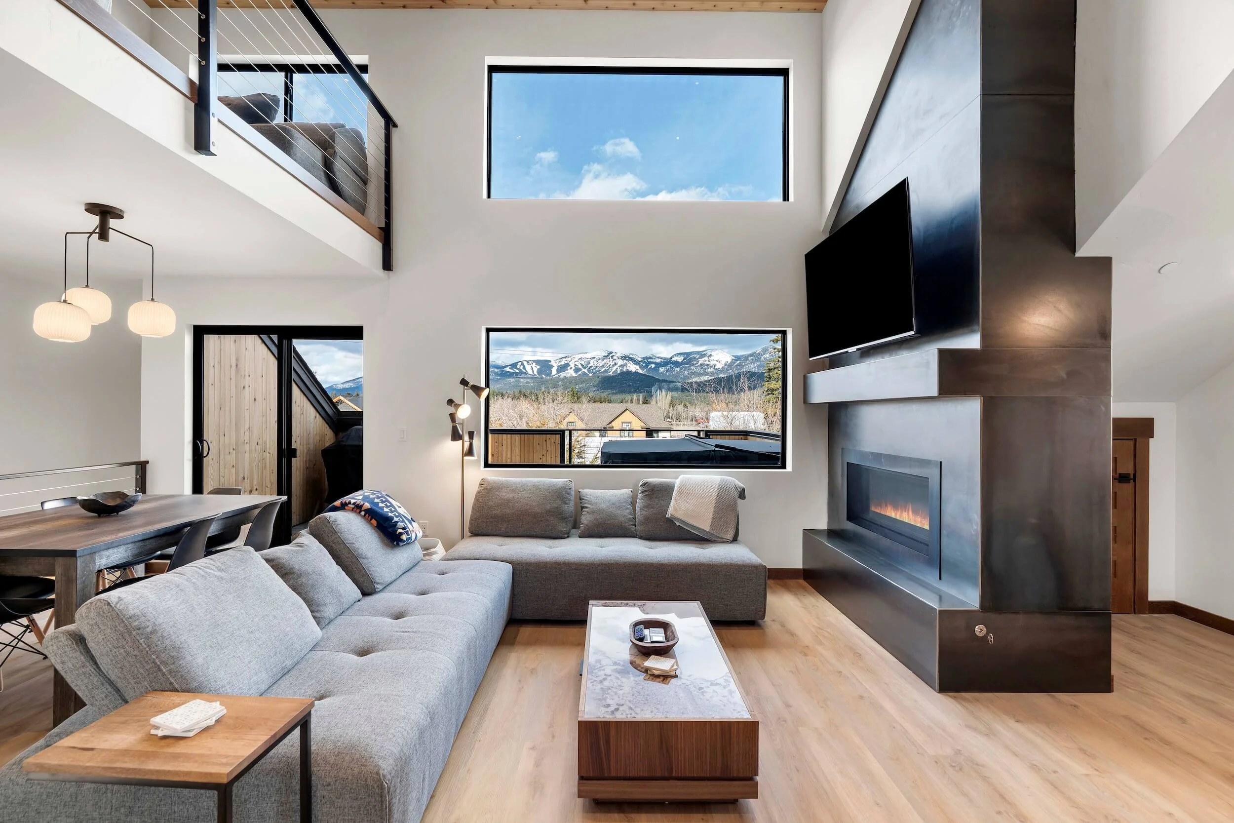 Modern living room with large windows showing mountain view, gray sectional sofa, fireplace, TV, wooden furniture, and ceiling lights.