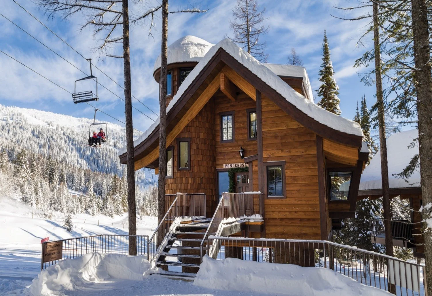 A wooden chalet-style house with snow on the roof, surrounded by snow-covered trees in a mountainous area, with ski lifts in the background and a sign that reads "Ponderosa" above the door.