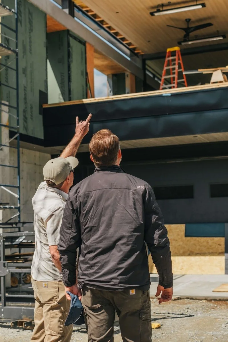 Two men working at a construction site, one explaining or pointing at a building under construction with exposed framing and a black exterior finish.