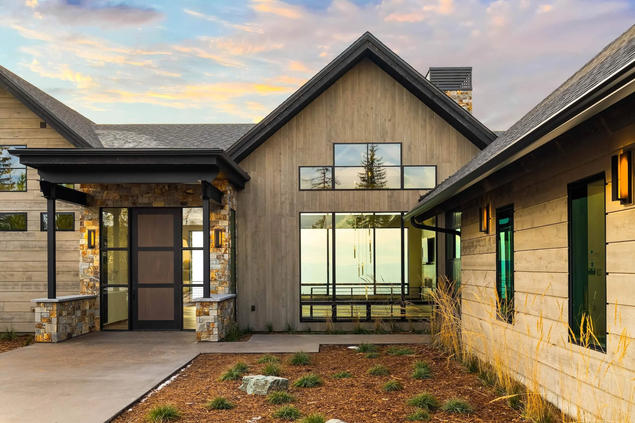 Modern house with wood and stone exterior, large glass windows, and a landscaped front yard at sunset.