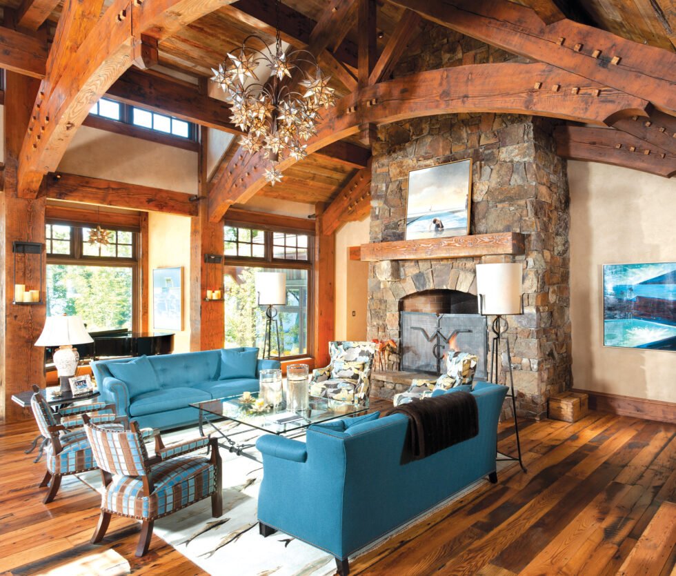 Living room with wooden beams, stone fireplace, blue sofas, plaid armchairs, large windows, and decorative lighting fixtures.