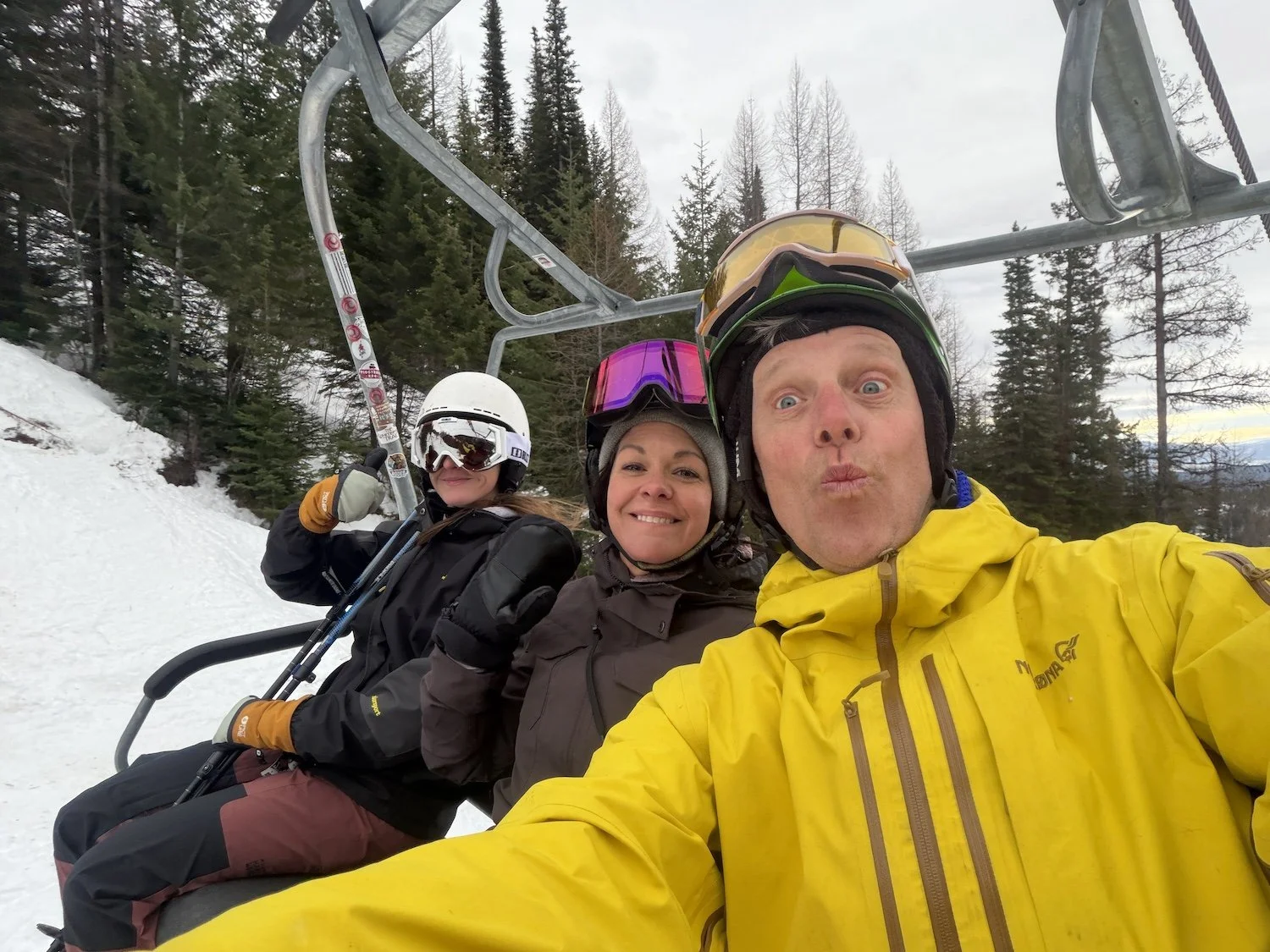 Three people riding a ski lift on a snowy mountain with pine trees in the background, wearing ski gear and helmets, taking a selfie.