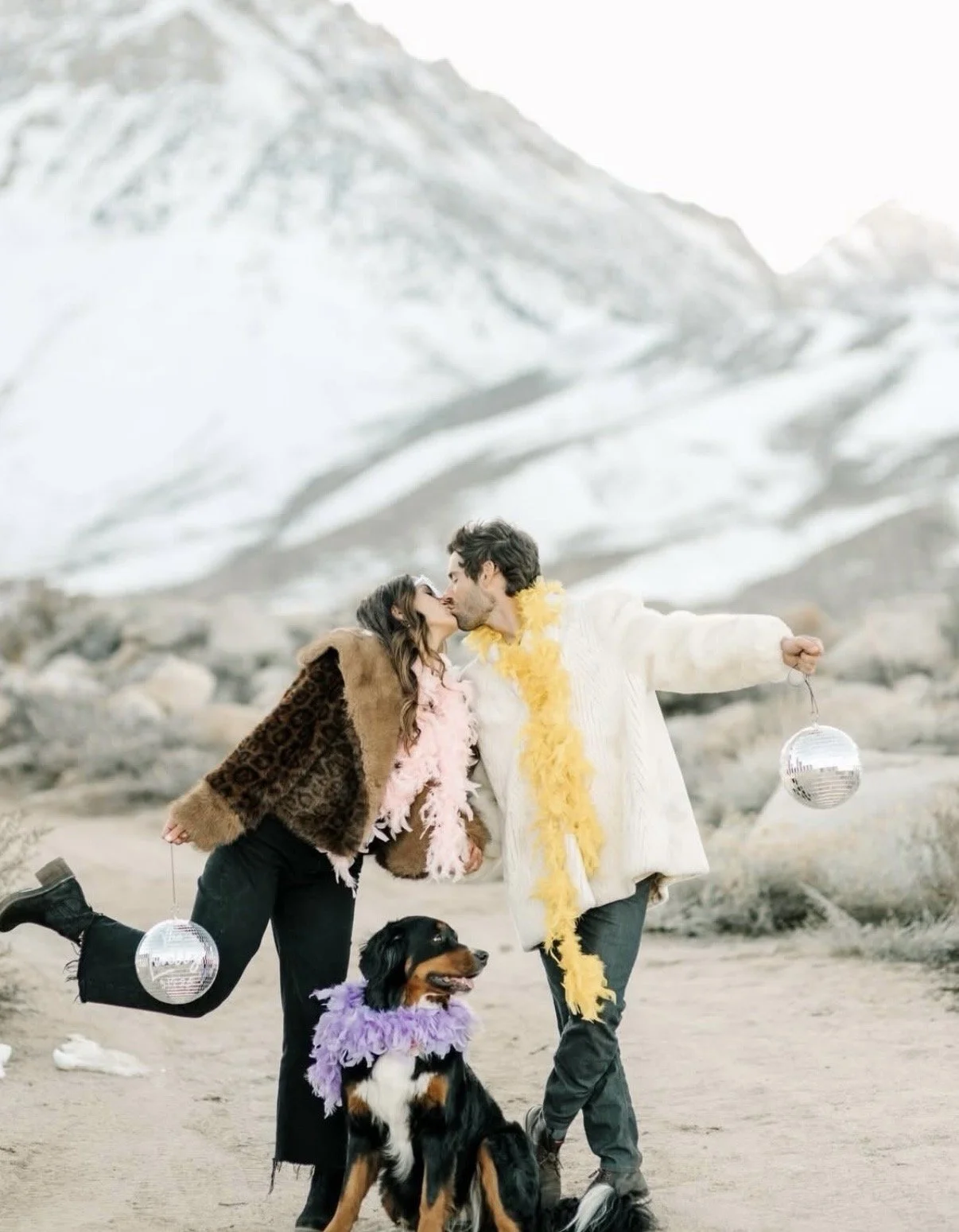 A couple kissing in a mountainous outdoor setting with a large mountain in the background, a dog wearing a purple feathered costume, and disco balls hanging from the people
