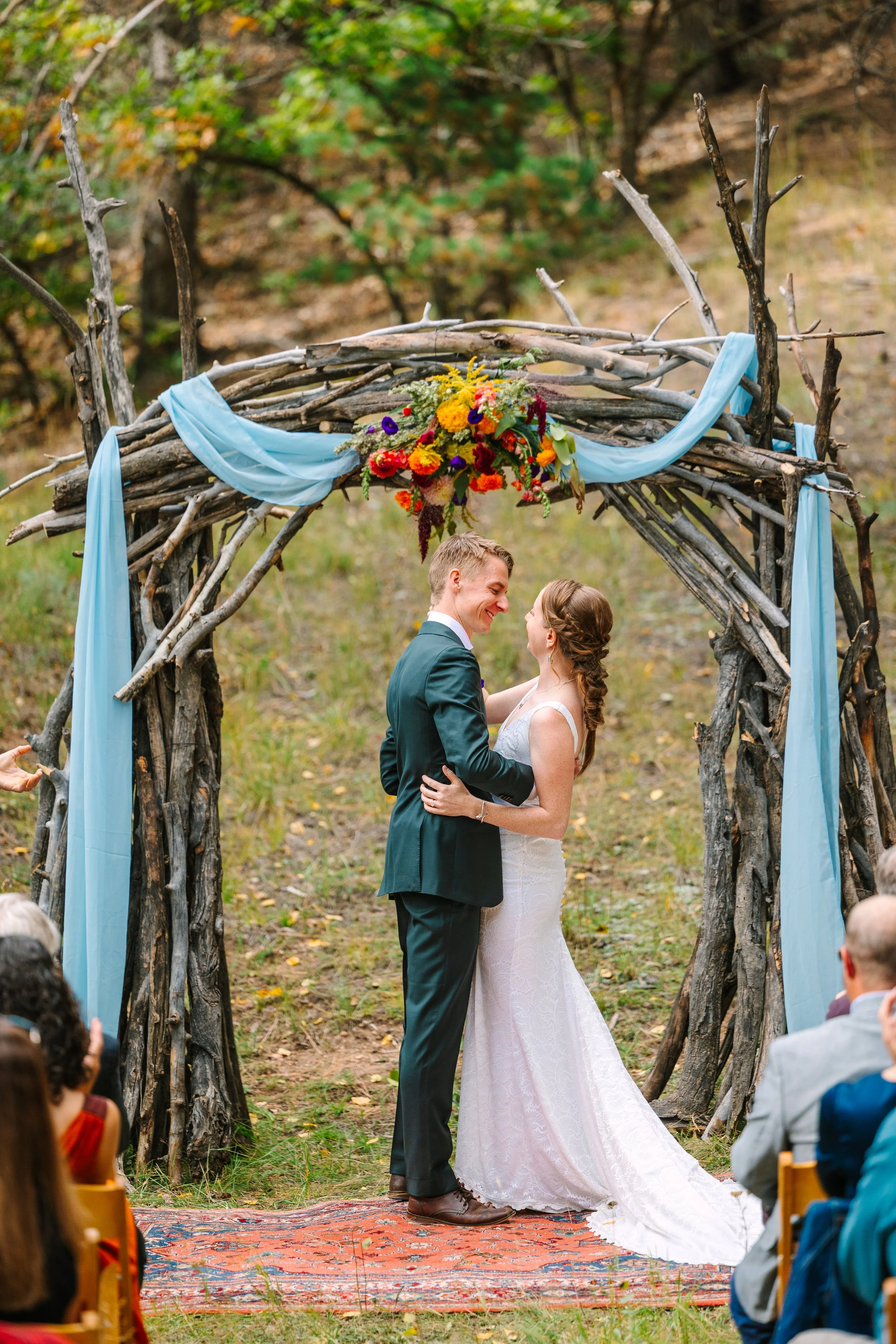 A bride and groom standing under a rustic arch made of branches, decorated with blue fabric and a colorful flower arrangement, during an outdoor wedding ceremony.