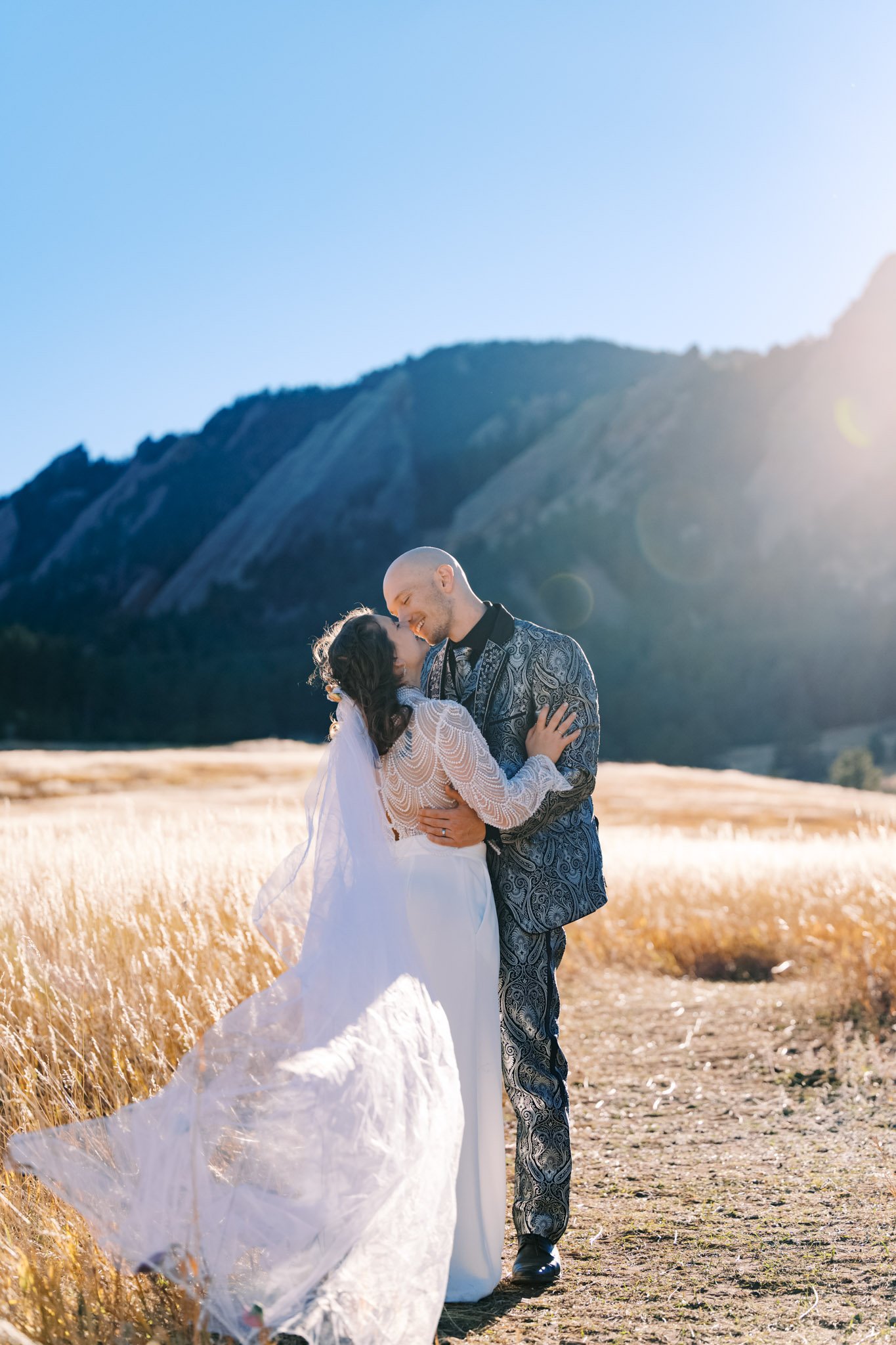 A couple is embracing in a field of tall, golden grass with mountains and a clear blue sky in the background, illuminated by sunlight.