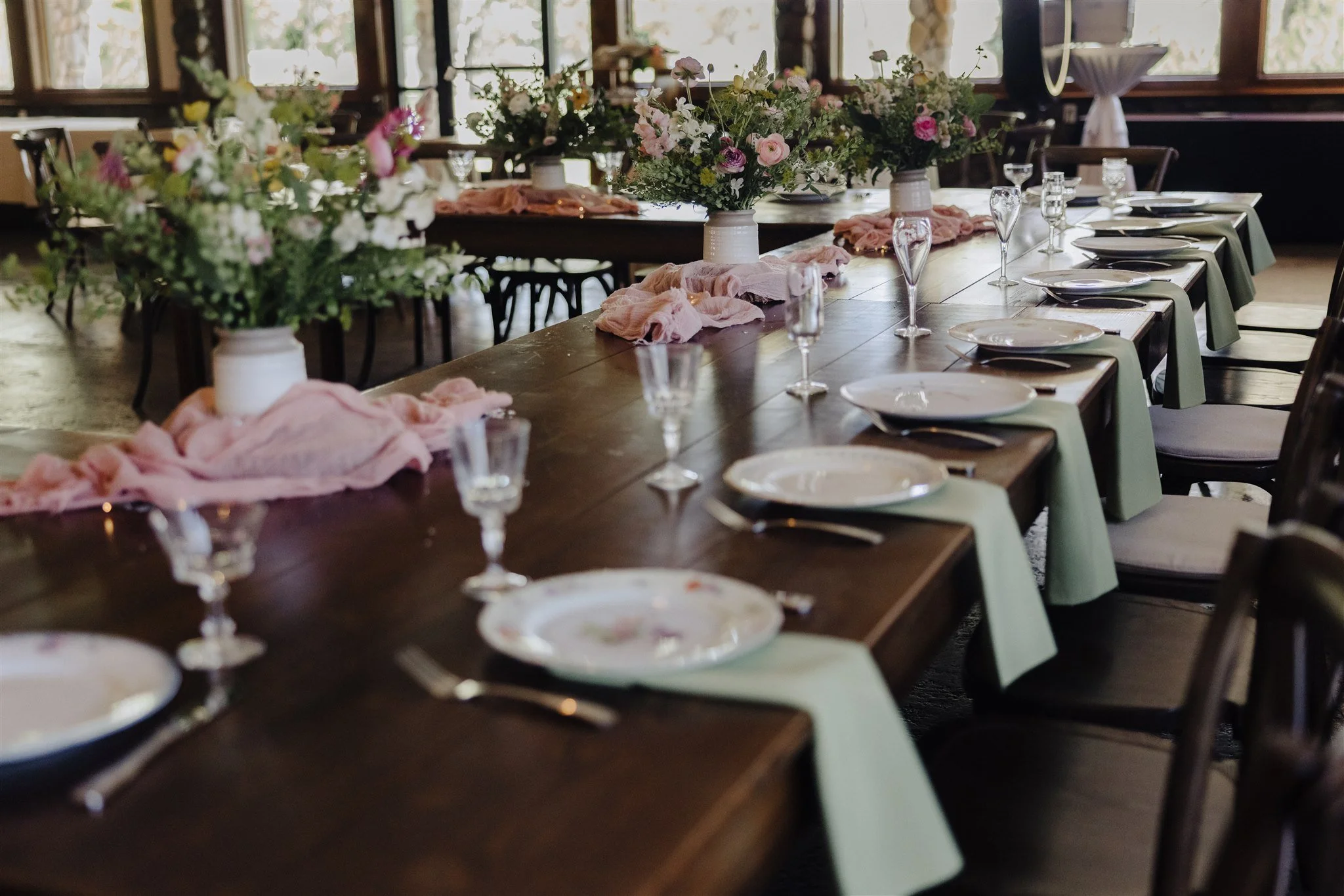 A long wooden dining table arranged for a special occasion with floral centerpieces in white vases, pink and white flowers, and pink cloth napkins. Set with white plates, clear wine glasses, and silverware. The room has large windows allowing natural light, and chairs placed around the table.