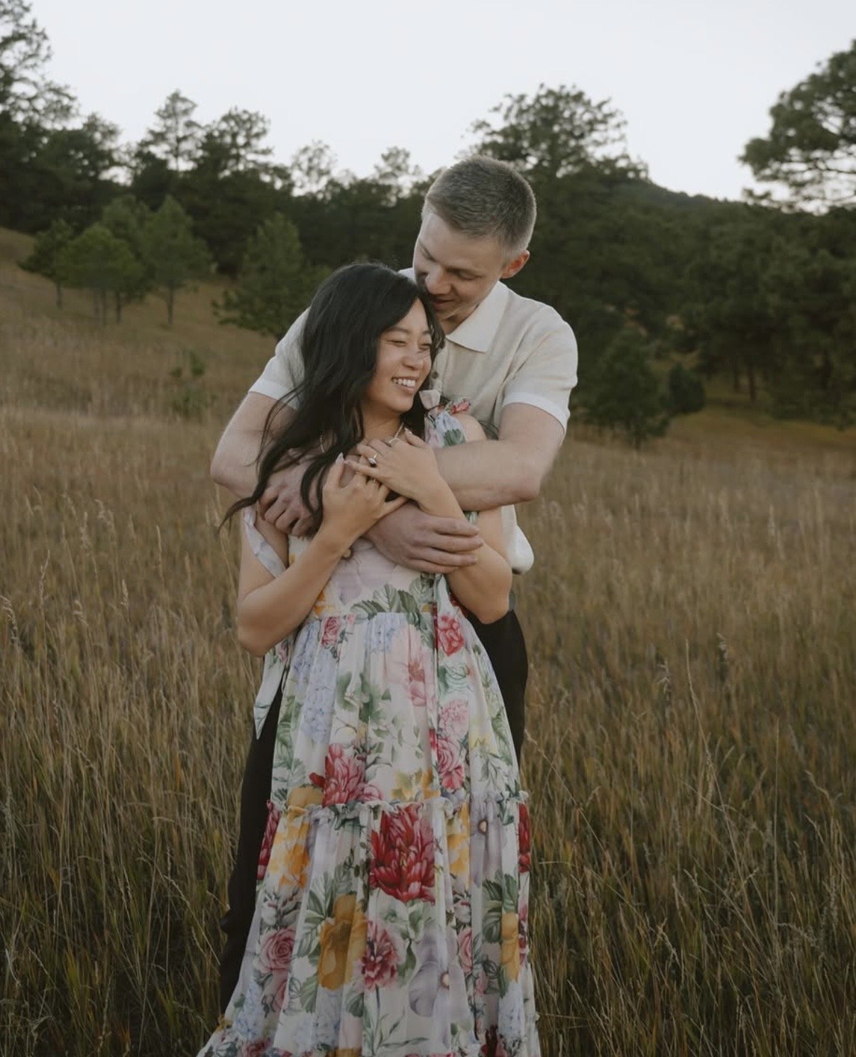 A couple hugging in a field of tall grasses with trees in the background during sunset.