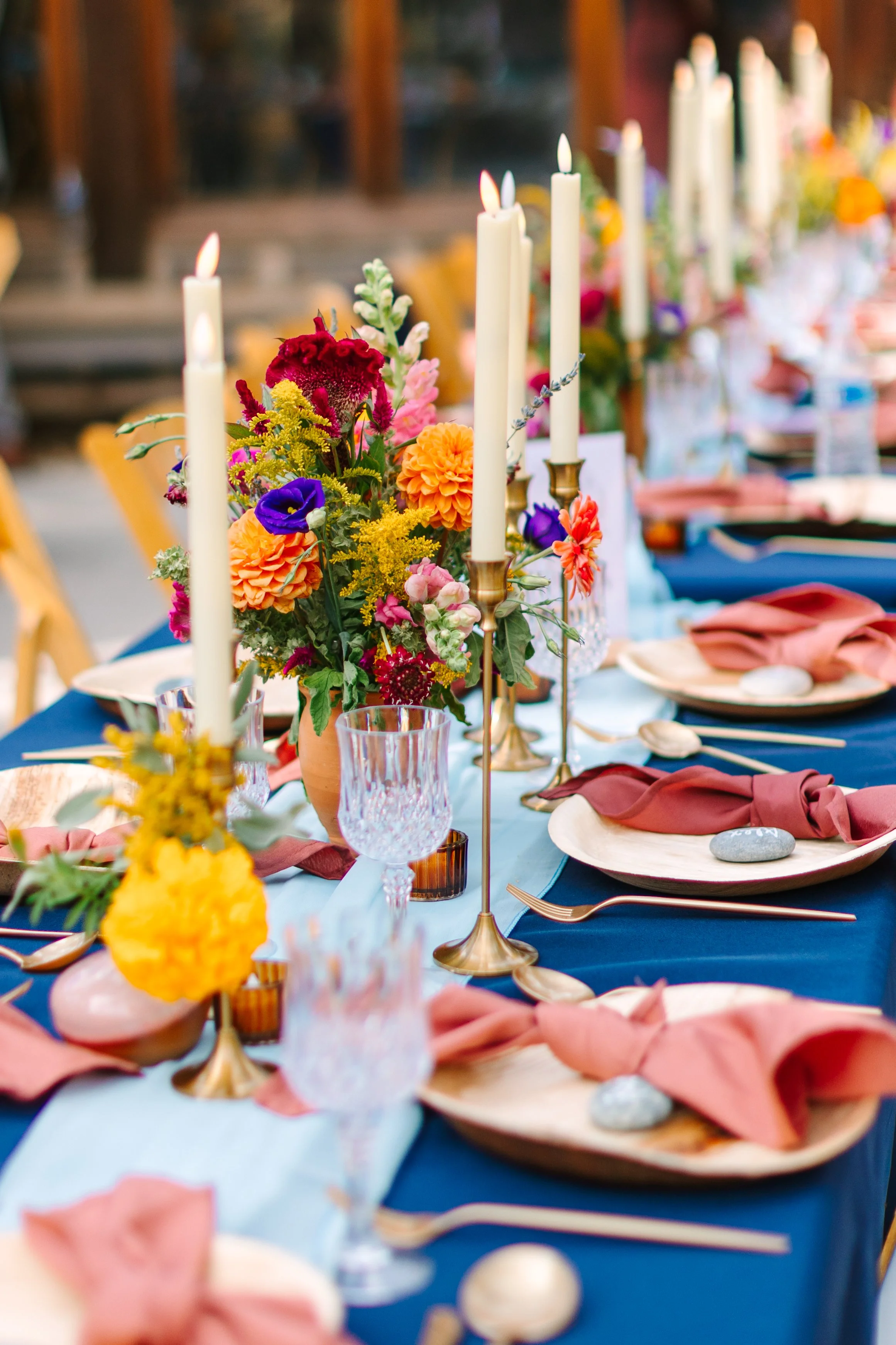 A beautifully decorated party table with tall white candles in gold holders, colorful fresh flower arrangements, pink napkins, and clear glassware on a blue tablecloth, set in an outdoor or rustic setting.