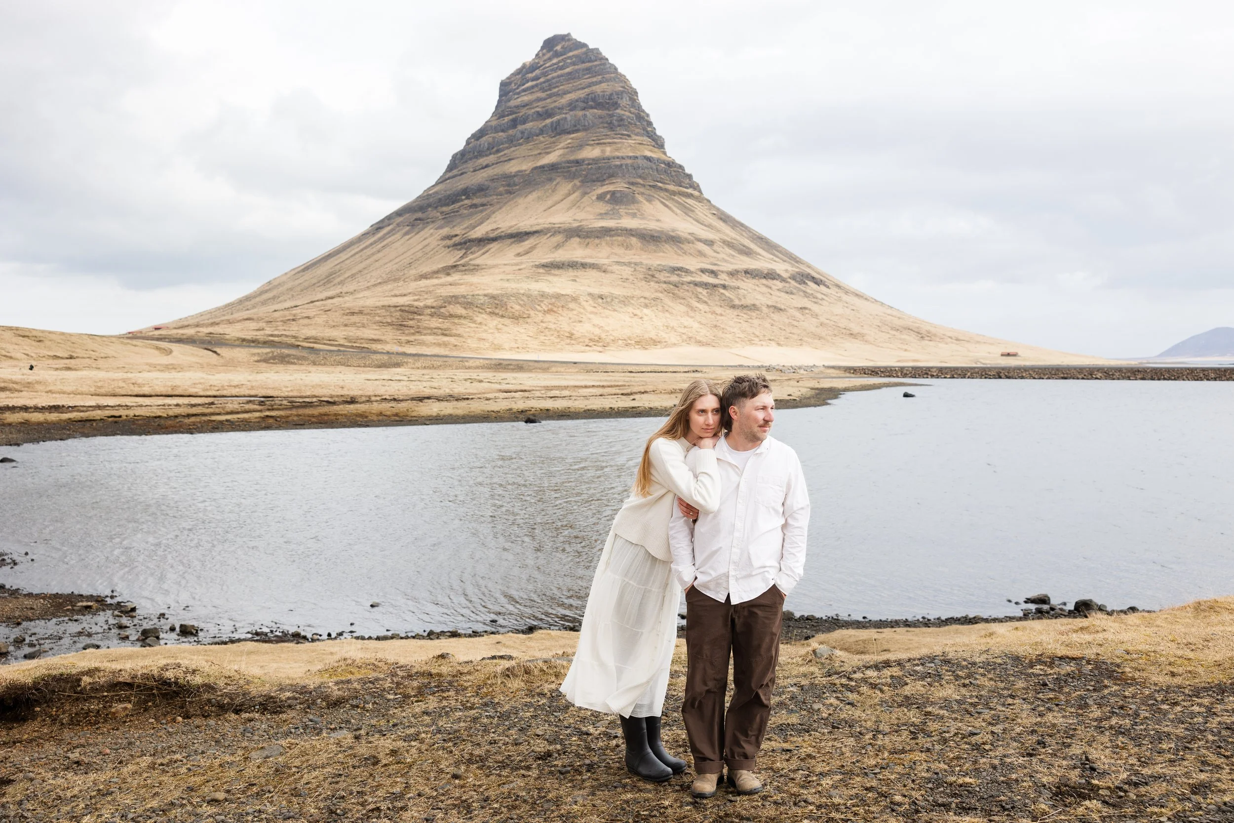 A young woman and man standing close together near a body of water with Kirkjufell mountain in the background, in a cold, cloudy outdoor landscape.