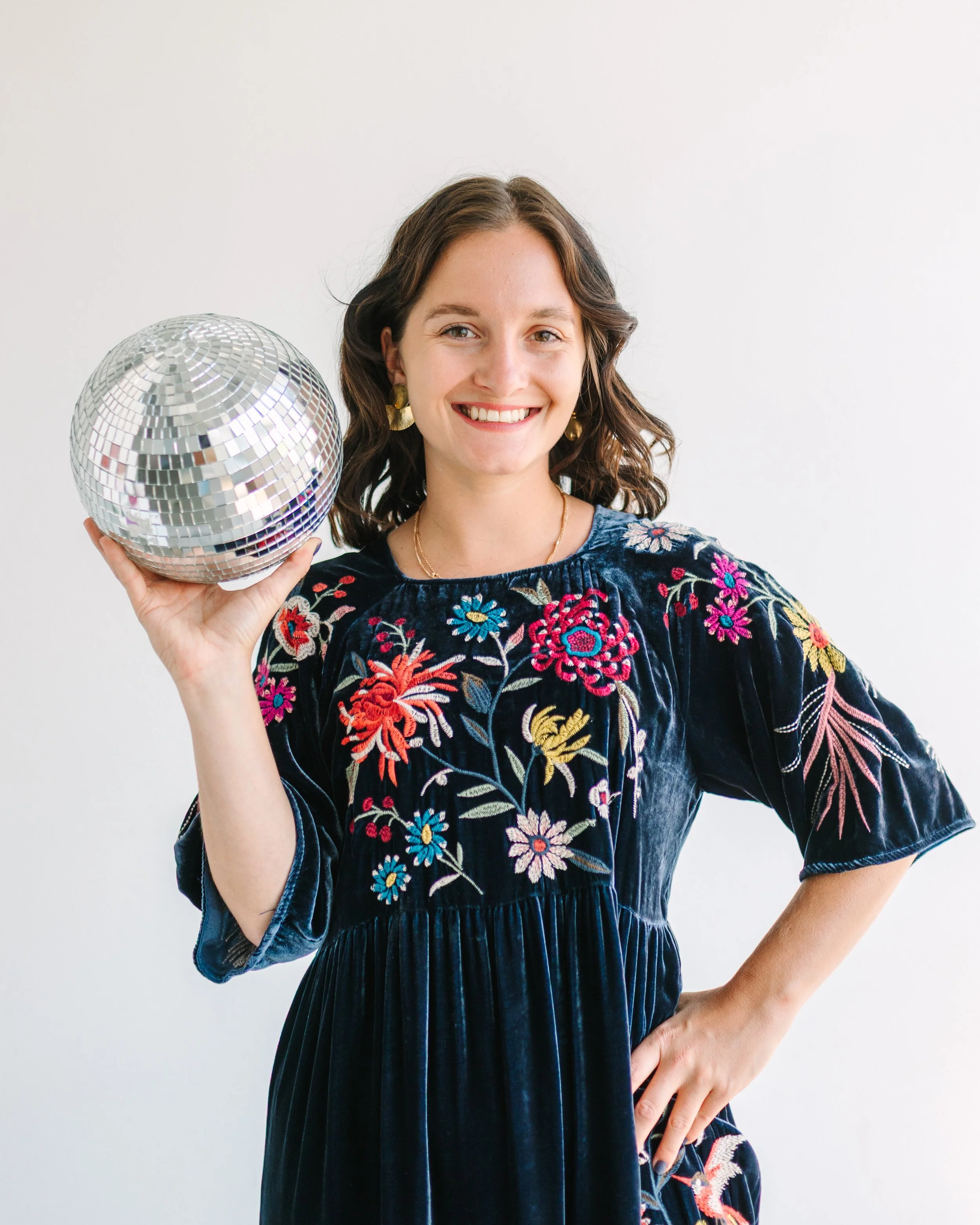 A smiling woman with shoulder-length wavy brown hair, wearing a dark blue embroidered dress with floral patterns, holding a disco ball in her right hand, standing against a plain white background.