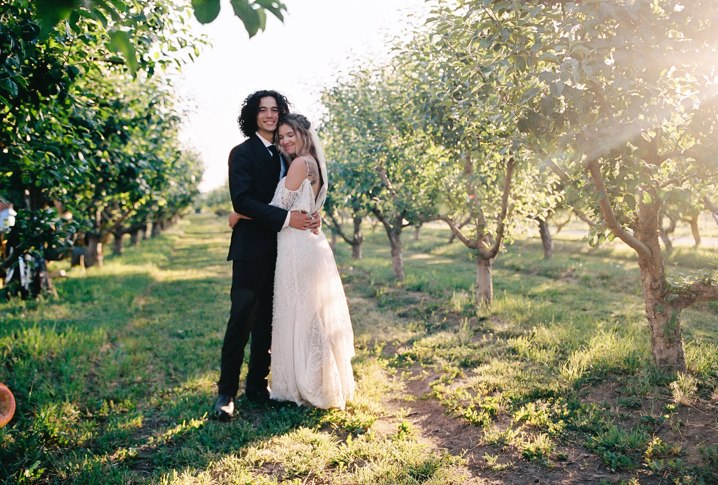 A smiling couple, dressed in wedding attire, embracing in a lush green orchard with sunlight filtering through the trees. Hotchkiss Colorado. Big Bs Orchard