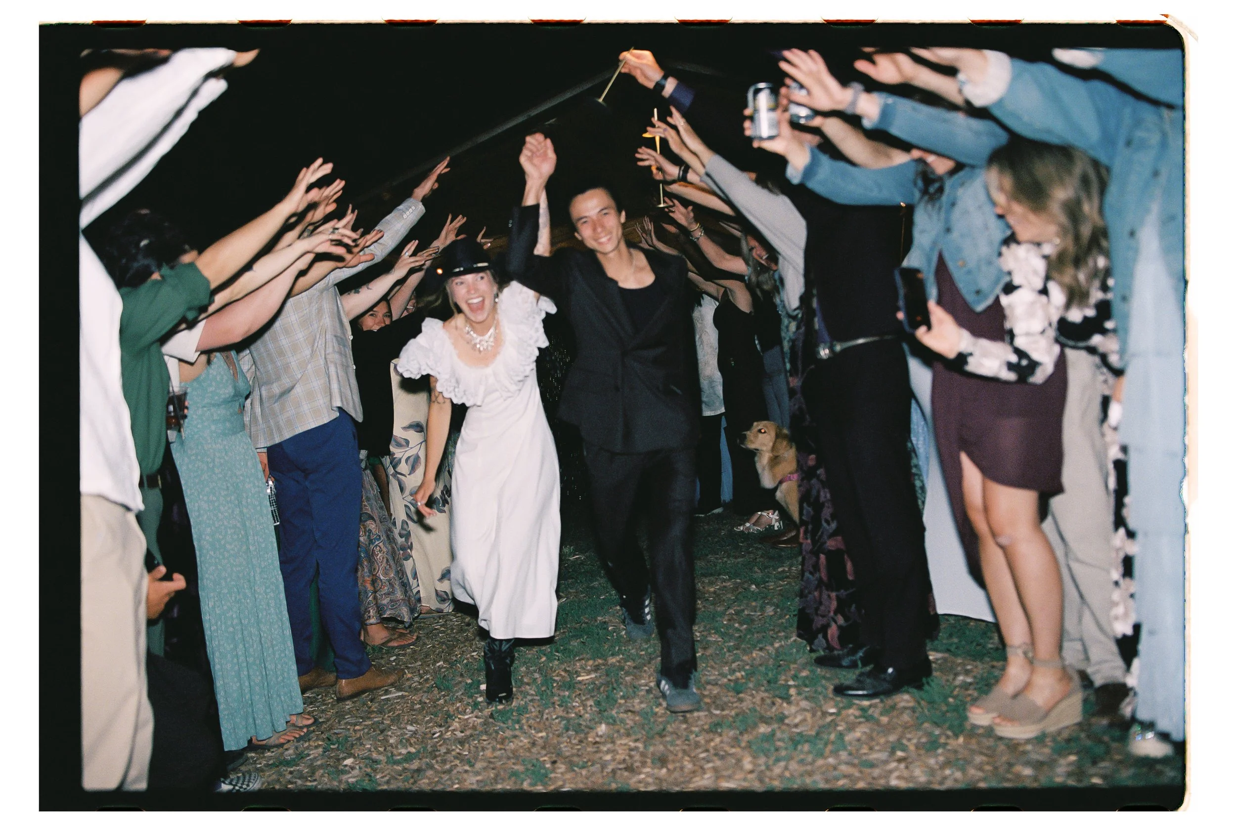 People walking through a tunnel formed by outstretched arms at an outdoor event, with a woman in a white dress and a man in black walking at the center.