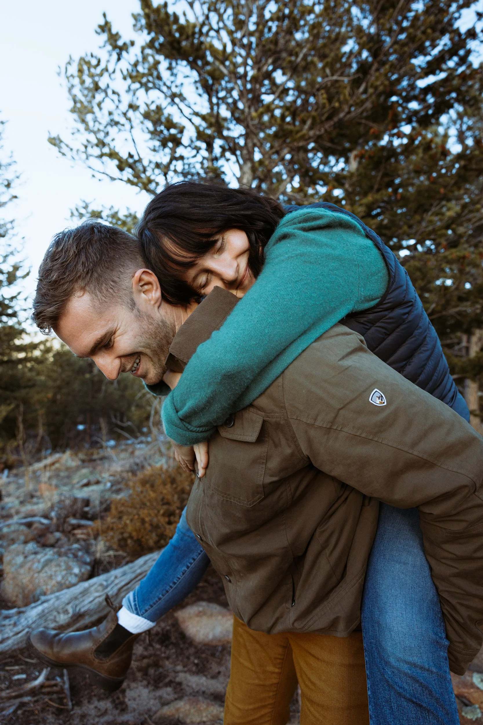 A couple outdoors, with the woman on the man's back, both smiling and enjoying a playful moment in nature with trees in the background.