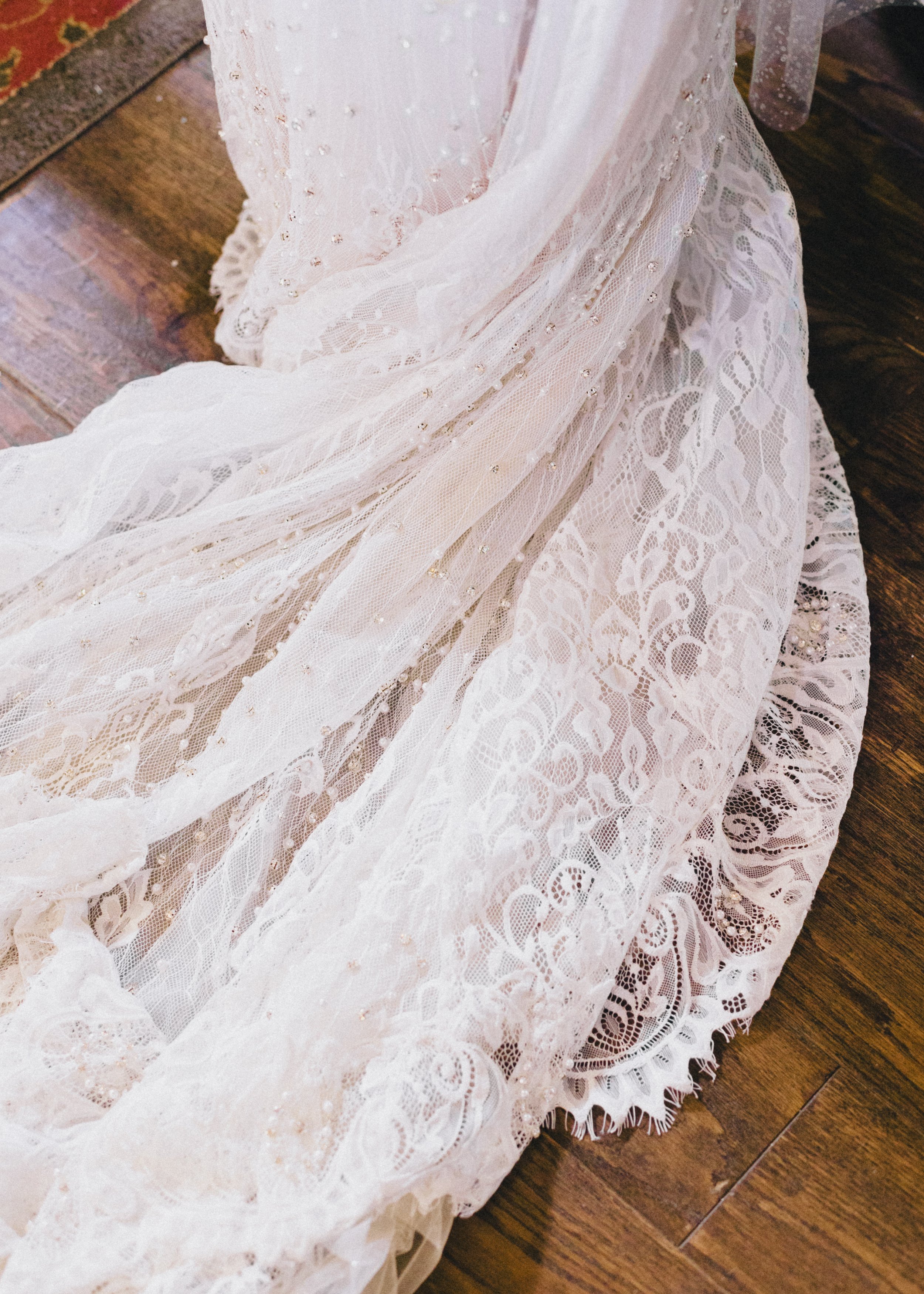 Close-up of a white lace wedding dress with intricate lace patterns and embellishments, draped over a wooden floor.