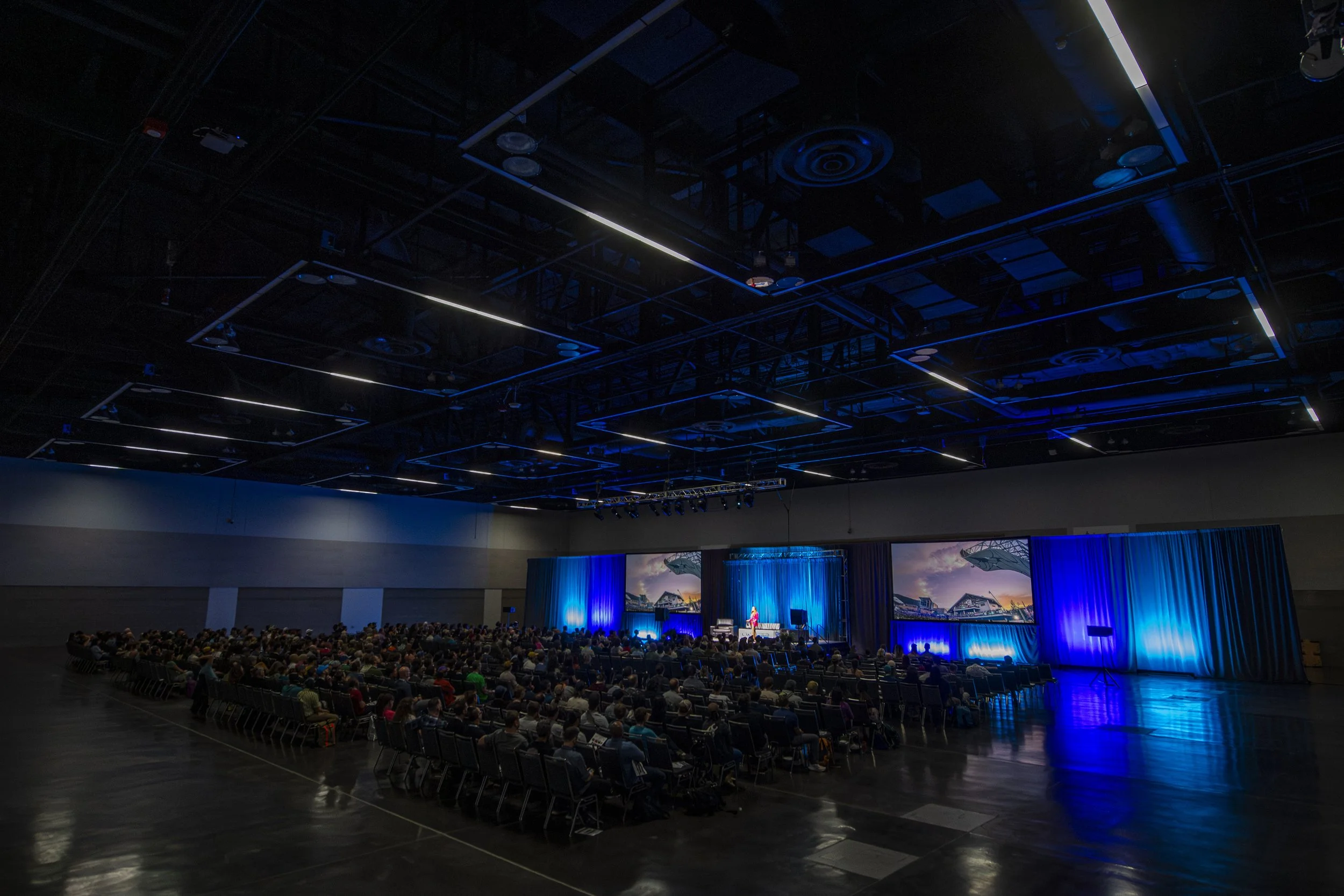 A large indoor auditorium filled with seated audience watching a presentation or performance on stage illuminated by blue lighting, with a large screen displaying images in the background. Climbing Wall Association Conference. Salt Lake City, Utah