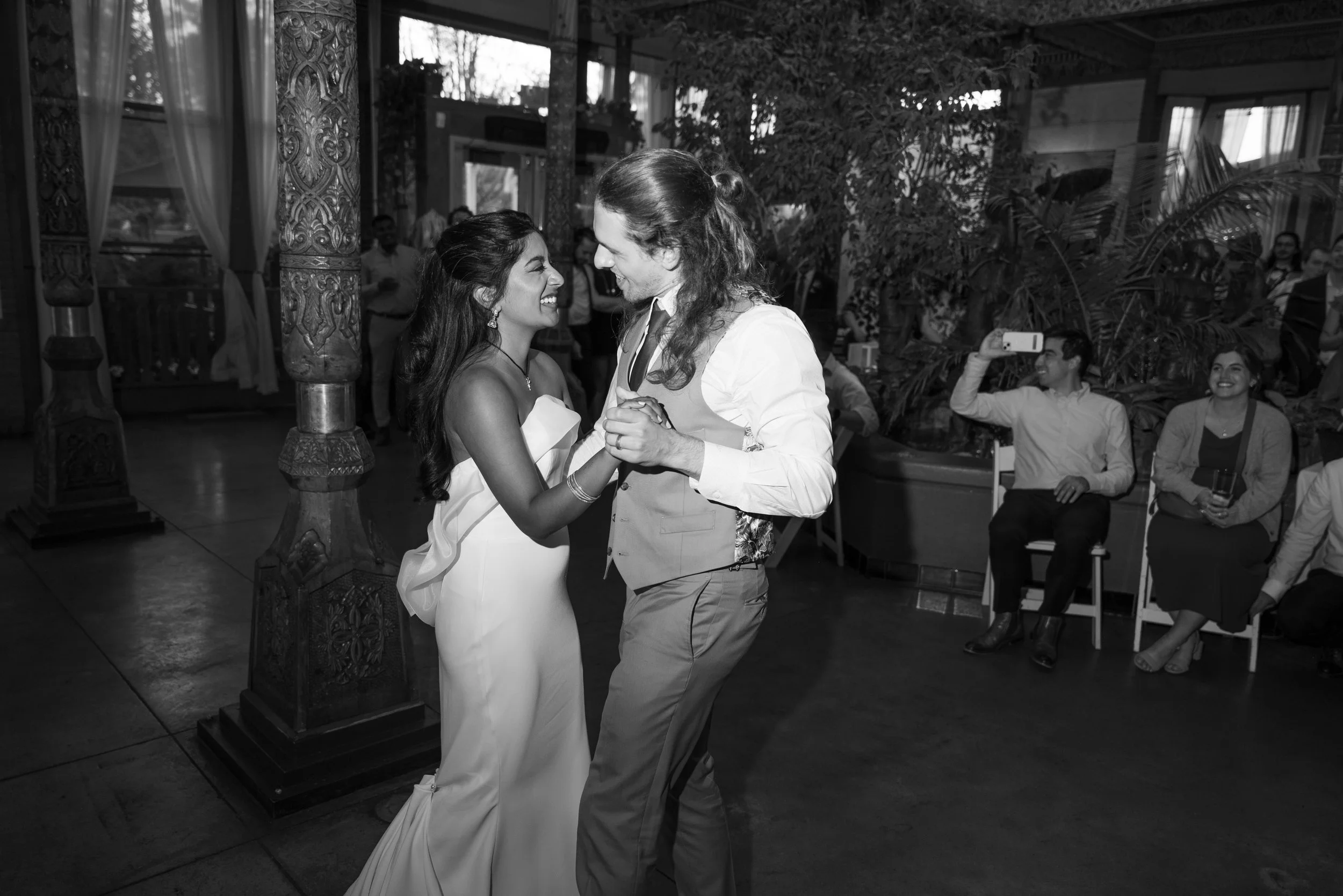 A couple dancing together at a wedding reception, smiling and gazing into each other's eyes, with guests seated and taking photos in the background.