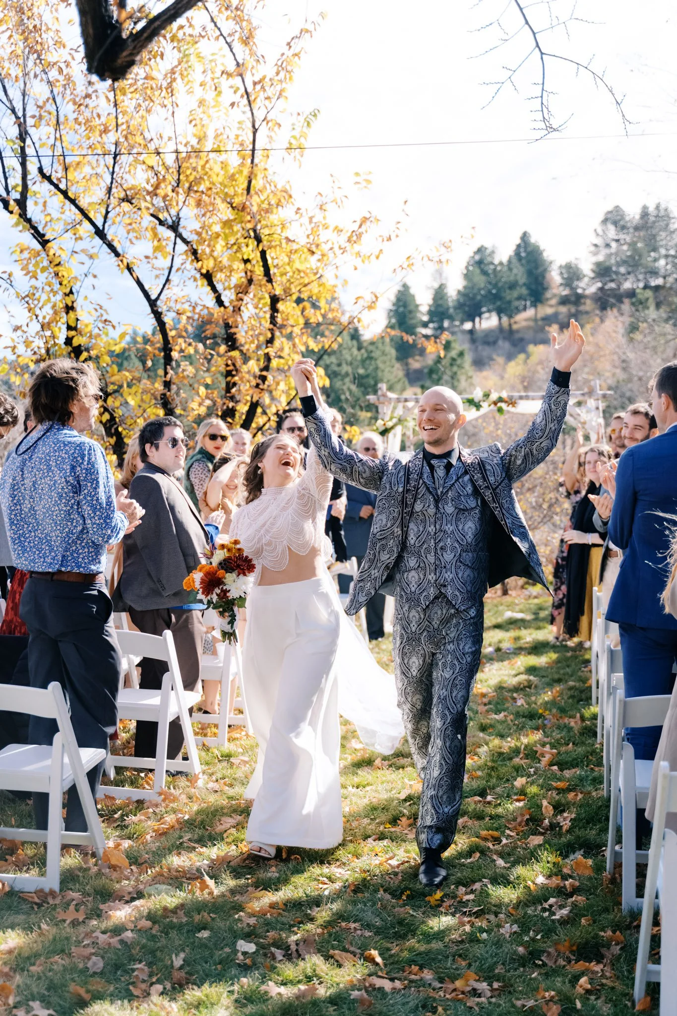 A joyful bride and groom celebrating their wedding outdoors with friends and family on a sunny autumn day, surrounded by fallen leaves and colorful trees.