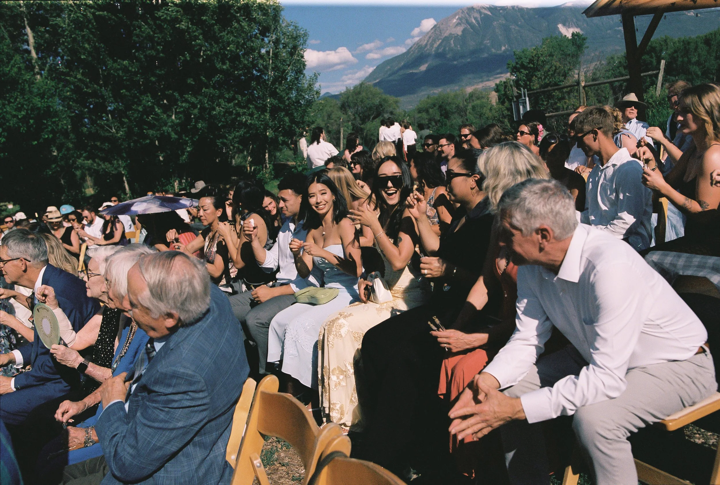 A group of people sitting outdoors on folding chairs during a sunny day, with green trees and mountains in the background, attending an event or gathering.