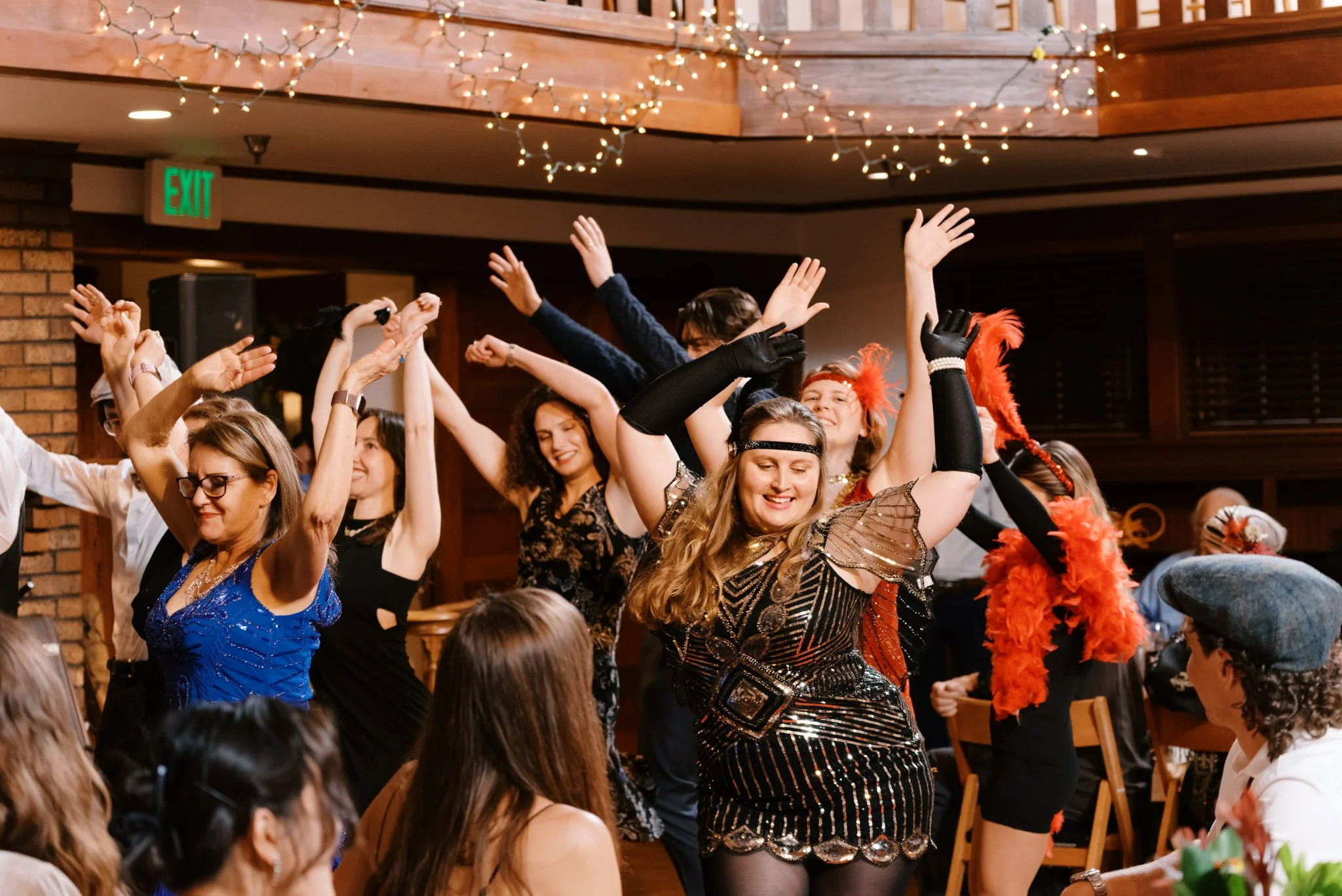People dancing and celebrating at a party, dressed in 1920s flapper costumes with feather accessories, in a decorated indoor venue with string lights.