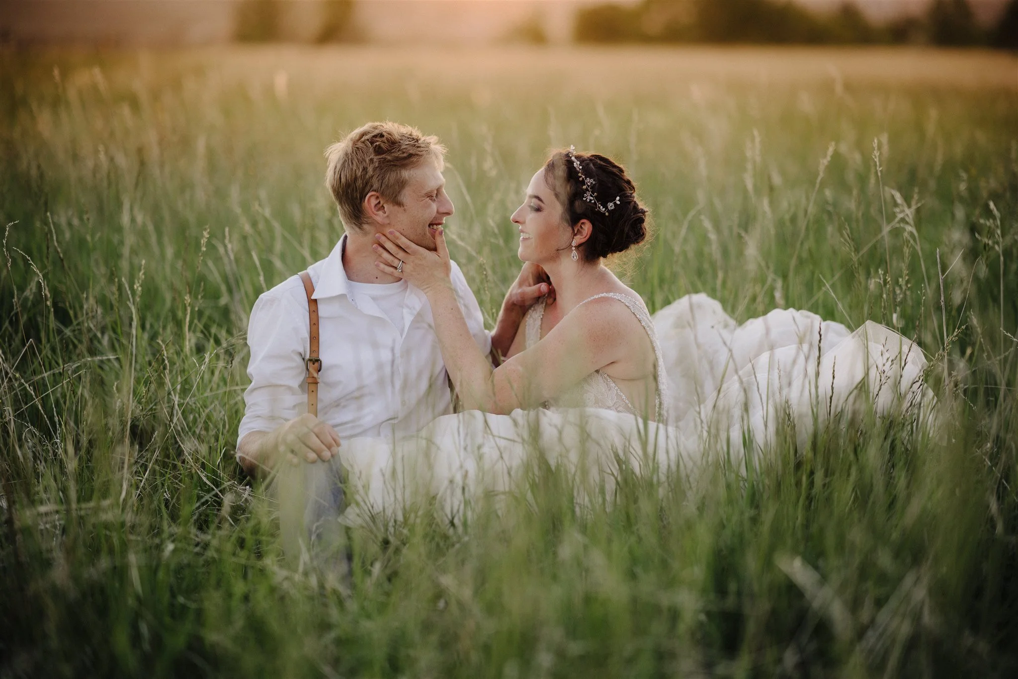 A couple sitting in a grassy field, dressed in wedding attire. The man is wearing a white shirt with suspenders, and the woman is wearing a wedding dress with a floral headpiece, smiling and touching each other's faces.