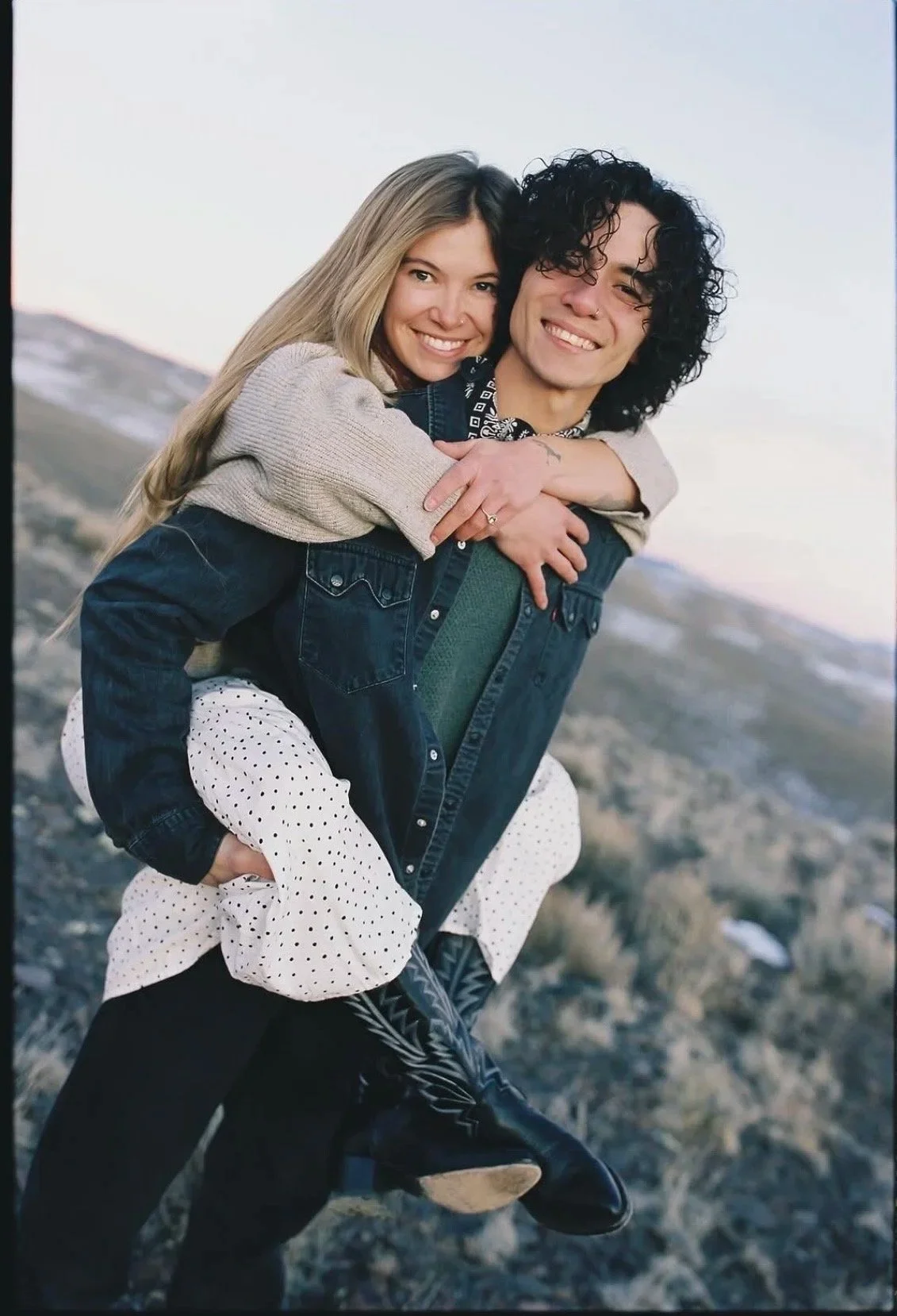 A young couple smiling and enjoying a piggyback ride outdoors in a rocky, open landscape during sunset or dusk.